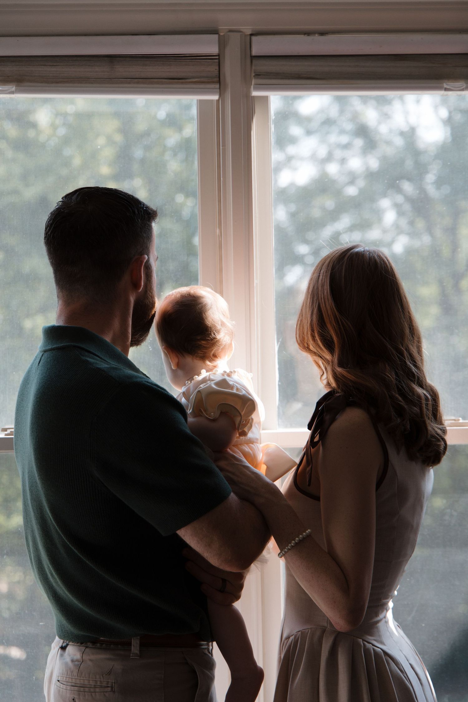 A couple holding their baby stands silhouetted against a bright window, gazing outside together.