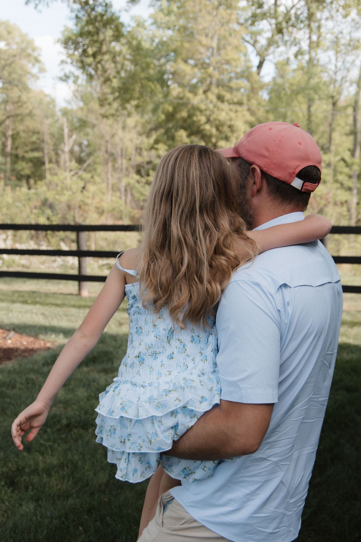 A man in a red cap holds a young girl in a floral dress outdoors near a fence with green trees. CHARLOTTE NC