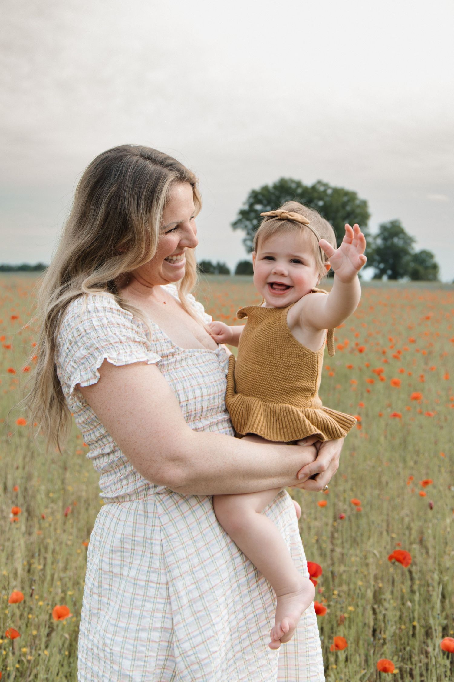 A smiling mother holds her laughing baby girl in a golden mustard outfit in a vibrant red poppy field. CHARLOTTE NC