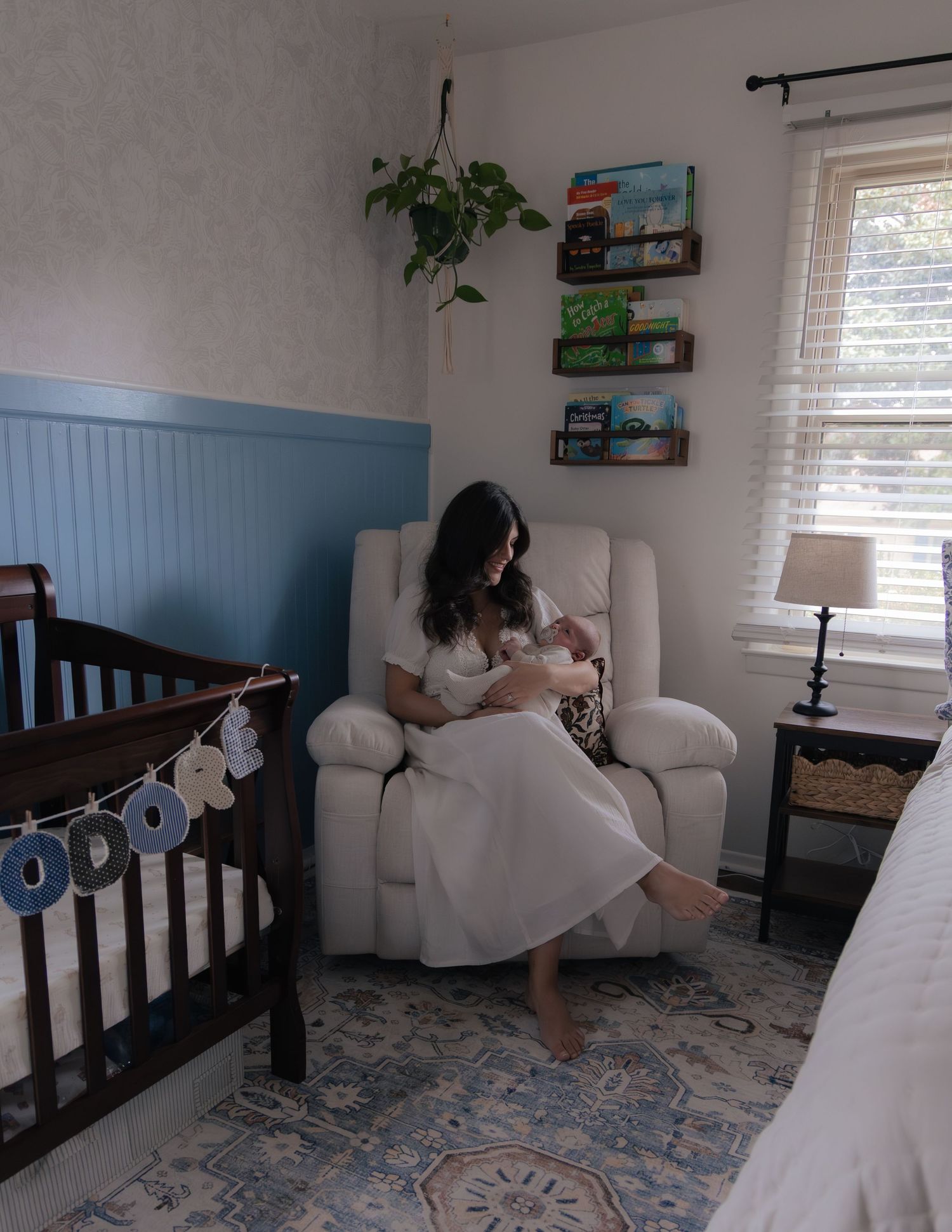 Mother in white dress nursing newborn in cream recliner in a blue and white nursery with crib and floating bookshelves. Charlotte NC