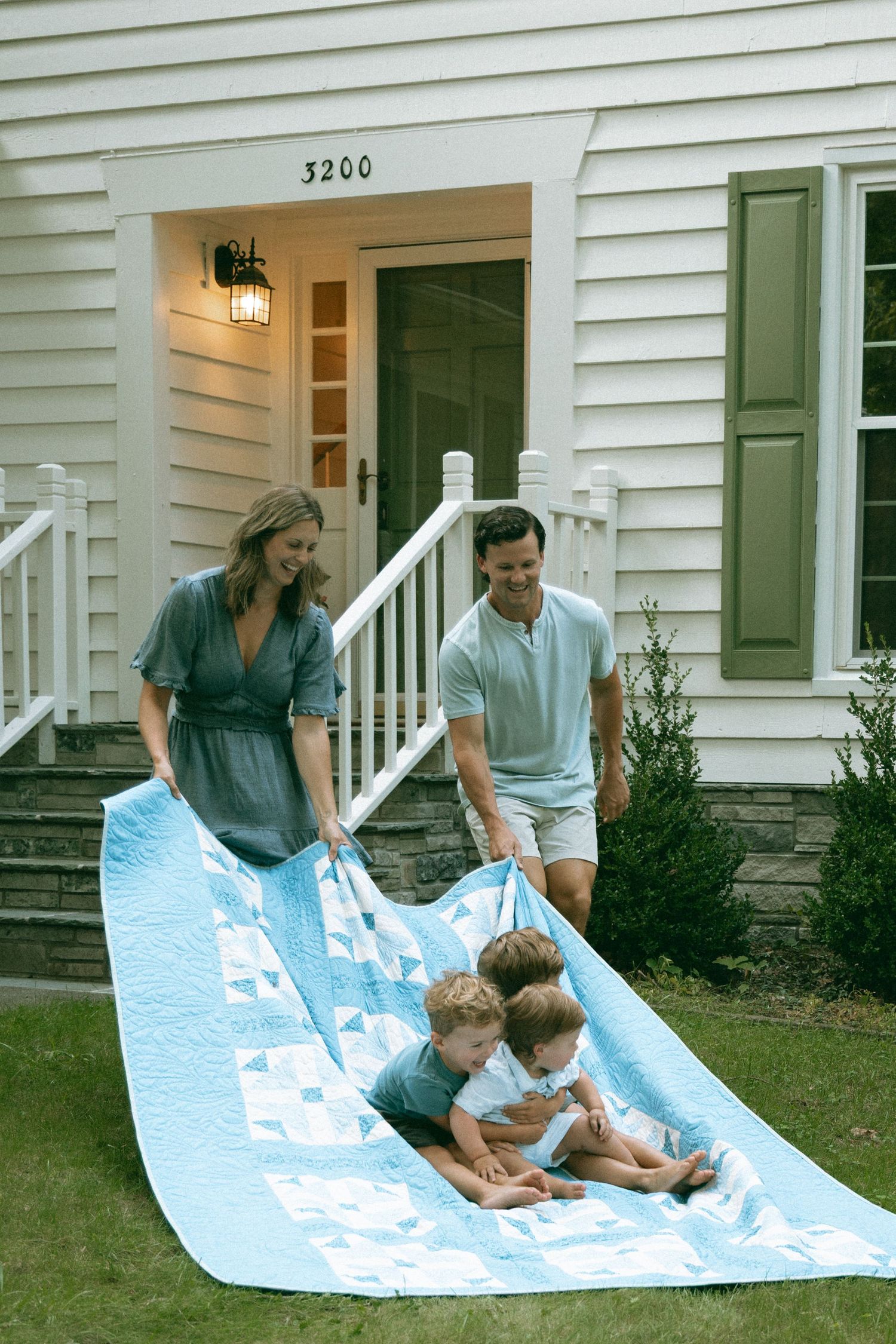 Family spreading a light blue quilt on green lawn outside white house numbered 3200 with green shutters.