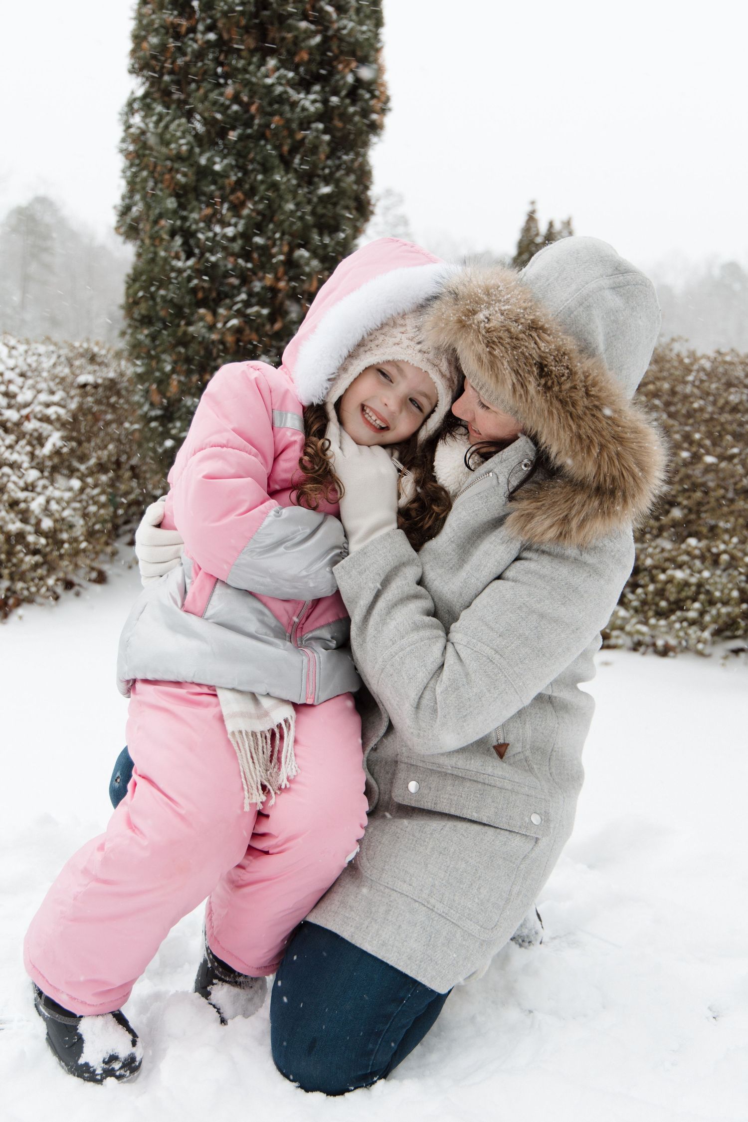 Mother and young daughter in pink snowsuit share a joyful winter embrace outdoors in snowy garden. Charlotte NC Matthews NC