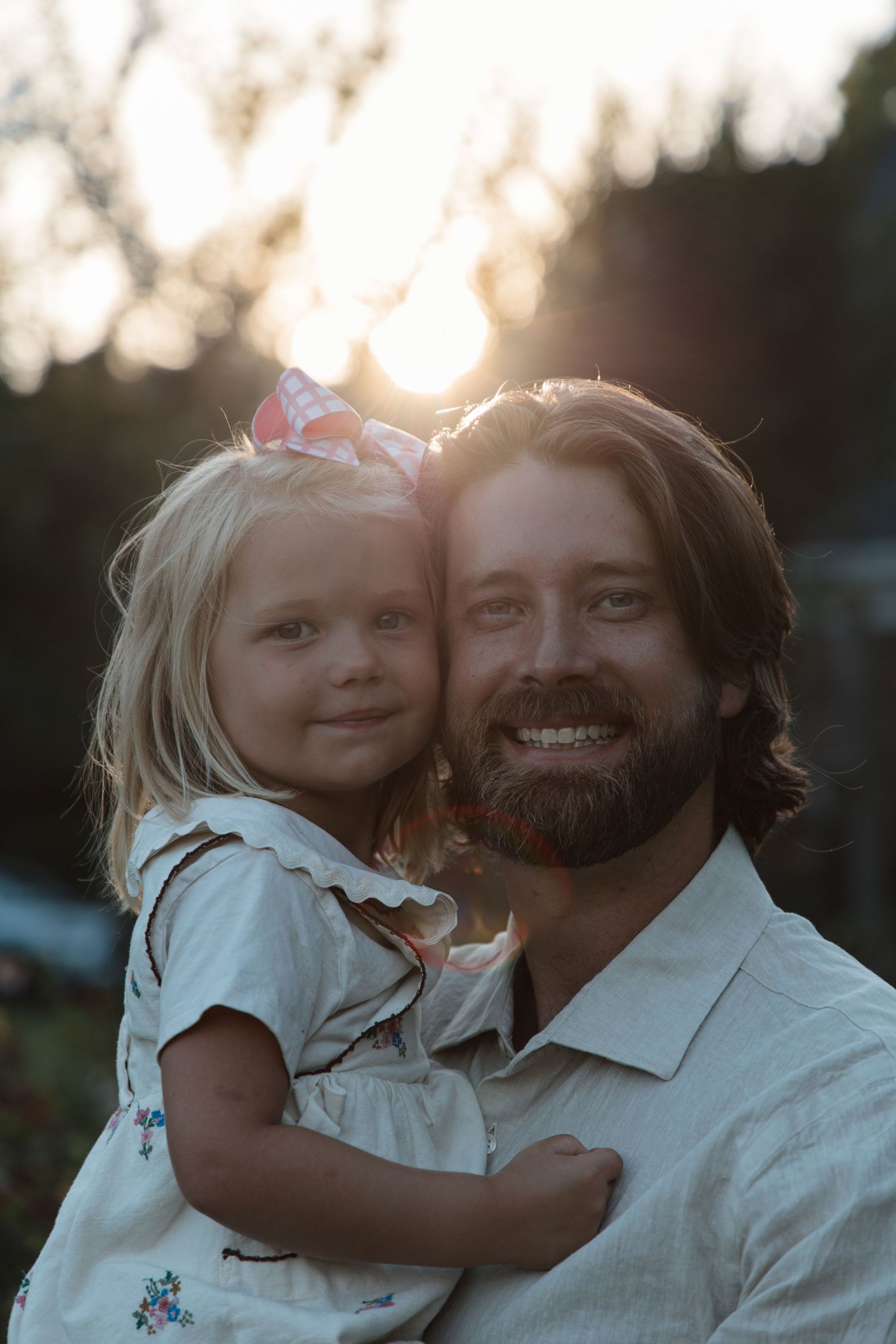 Smiling bearded man holding a young blonde girl with a bow at golden hour sunset outdoors.