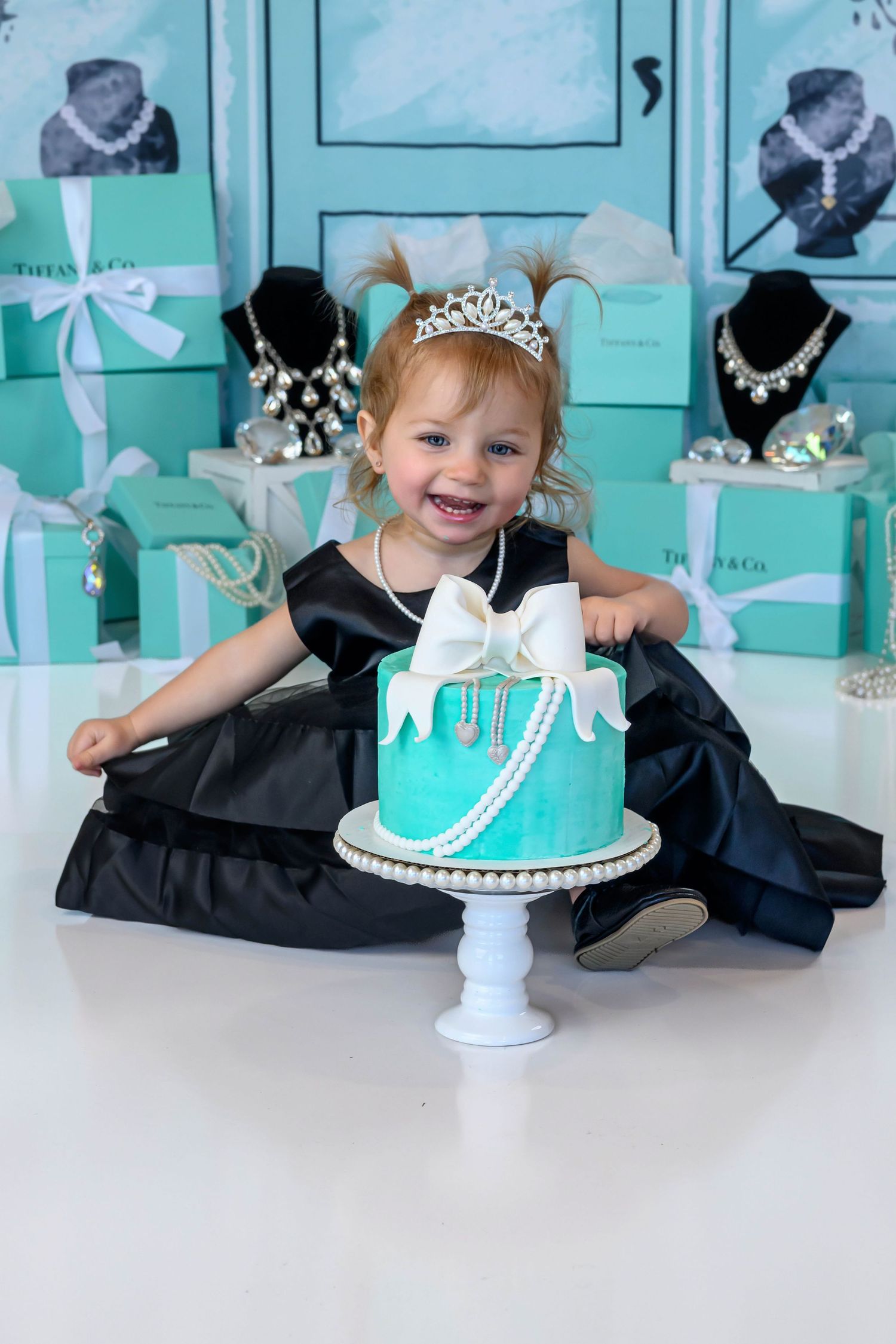 Smiling toddler girl in black dress and tiara sits with teal Tiffany-themed birthday cake and gift boxes.