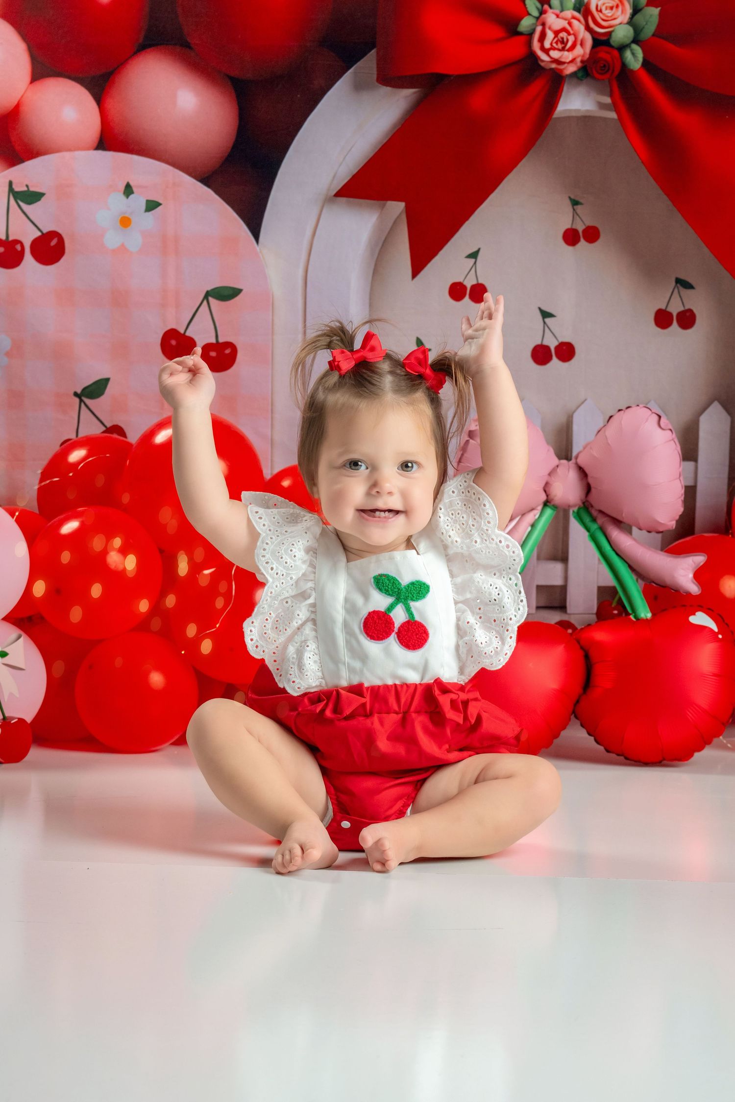 Smiling baby girl in cherry outfit sitting among red balloons and cherry decorations at birthday.