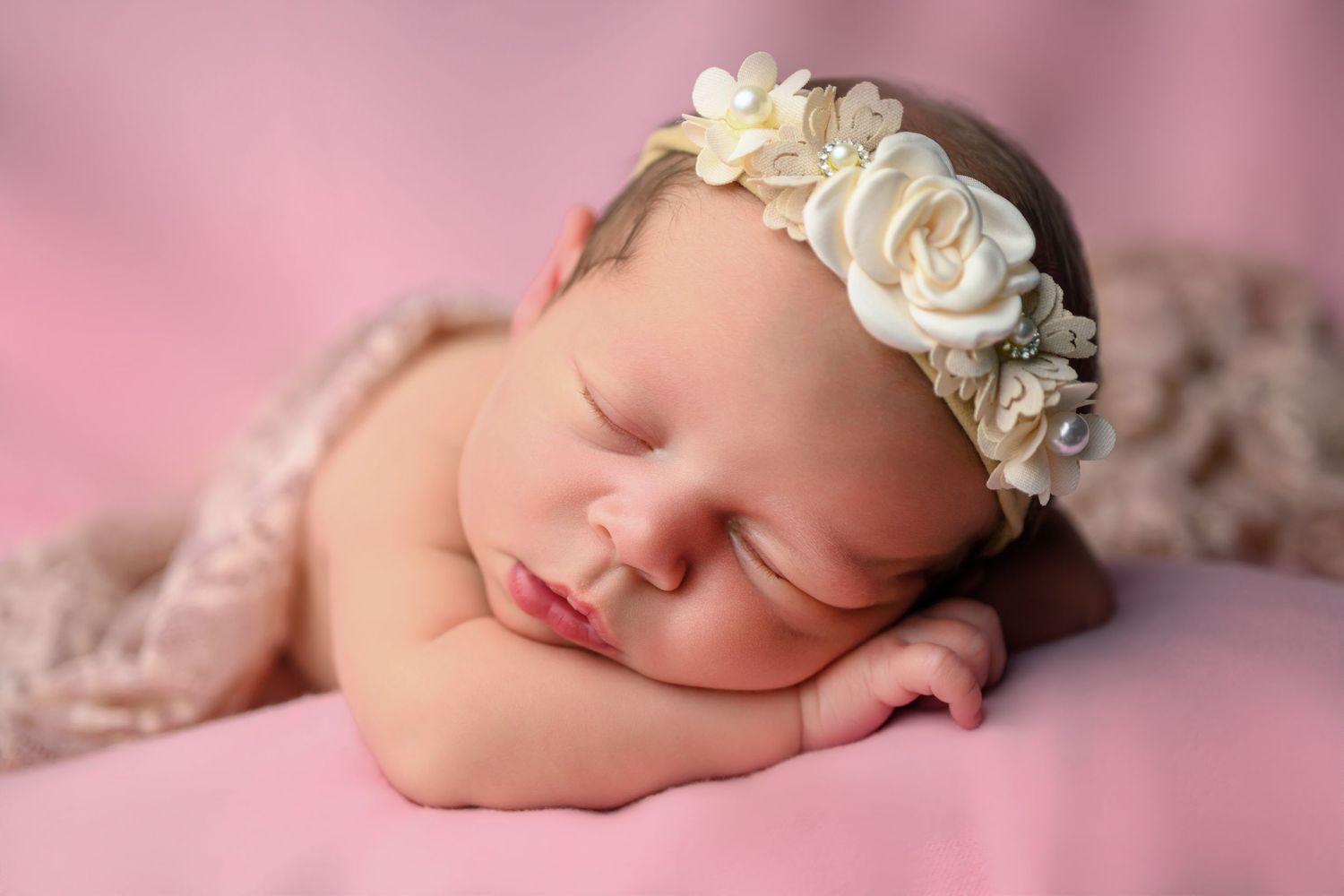 Peaceful newborn baby girl sleeping on pink blanket wearing a delicate cream floral headband.