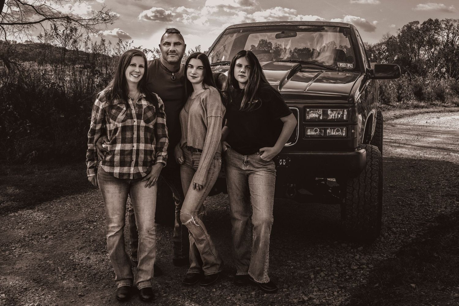 Sepia-toned family portrait of four people posing by a pickup truck on a rural dirt road.