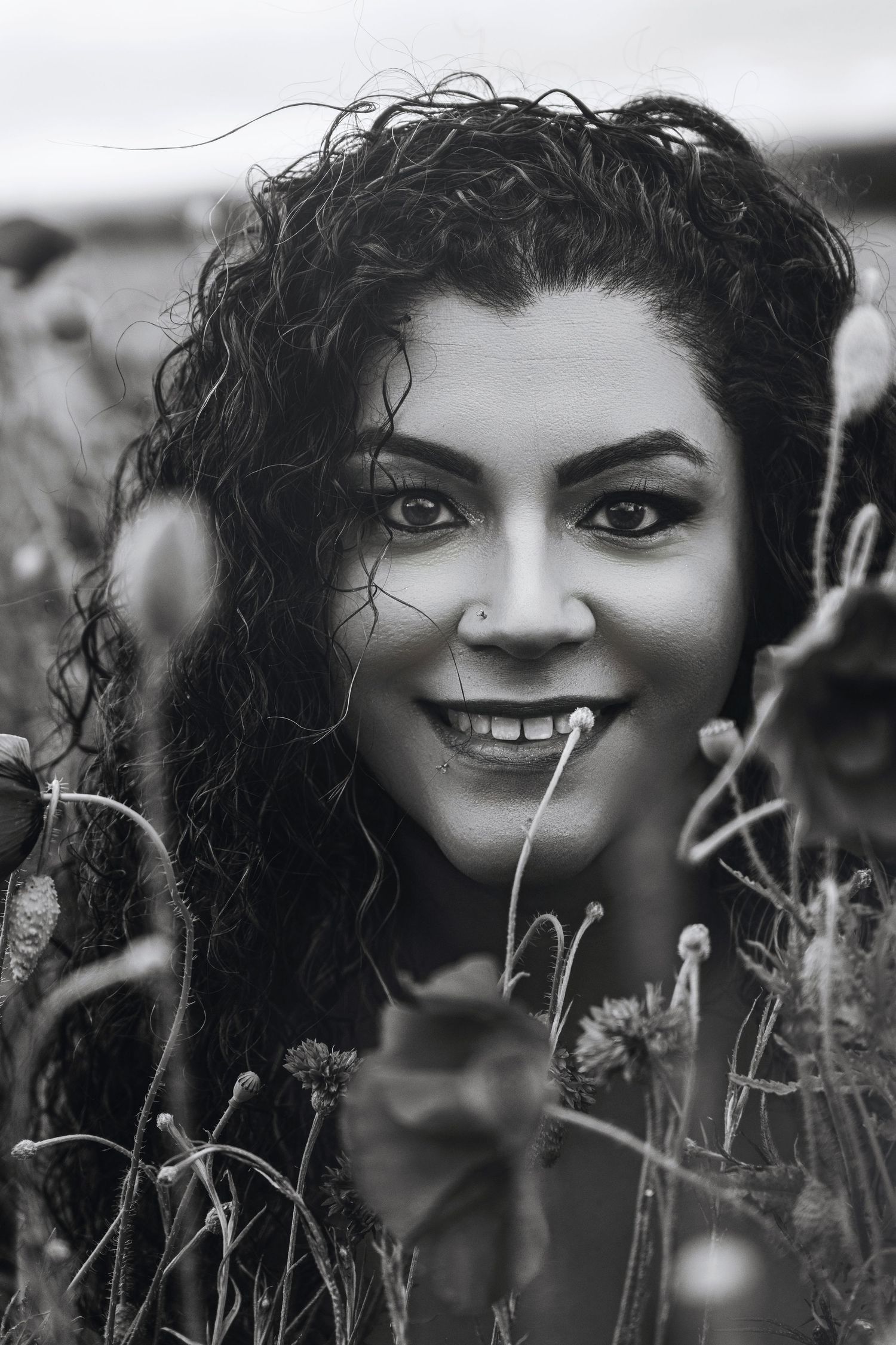 Black and white portrait of curly-haired woman smiling among wildflowers in an open field.