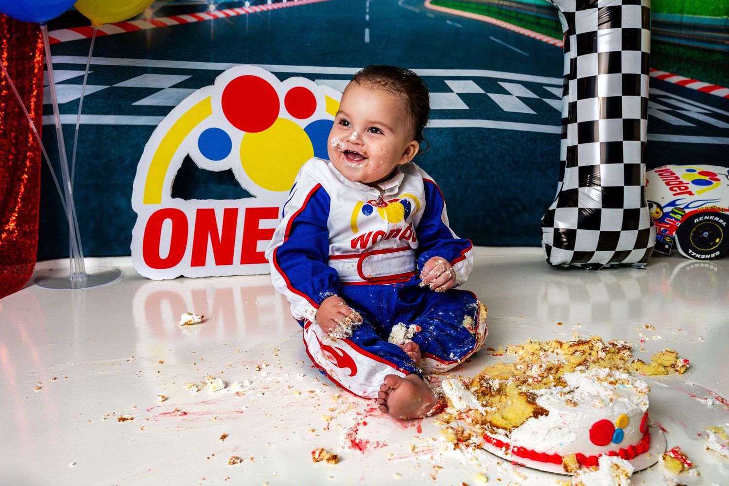 Happy baby in racing suit enjoys first birthday cake smash with colorful decorations.