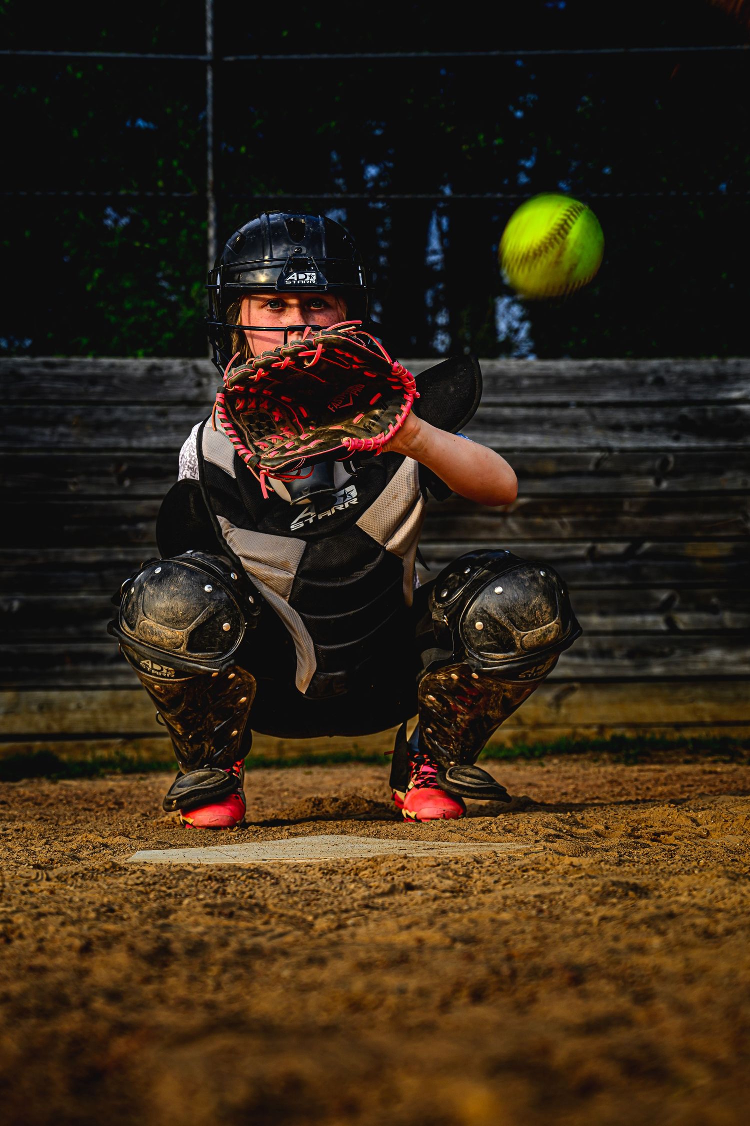 Youth softball catcher in full protective gear awaits incoming yellow softball at plate.