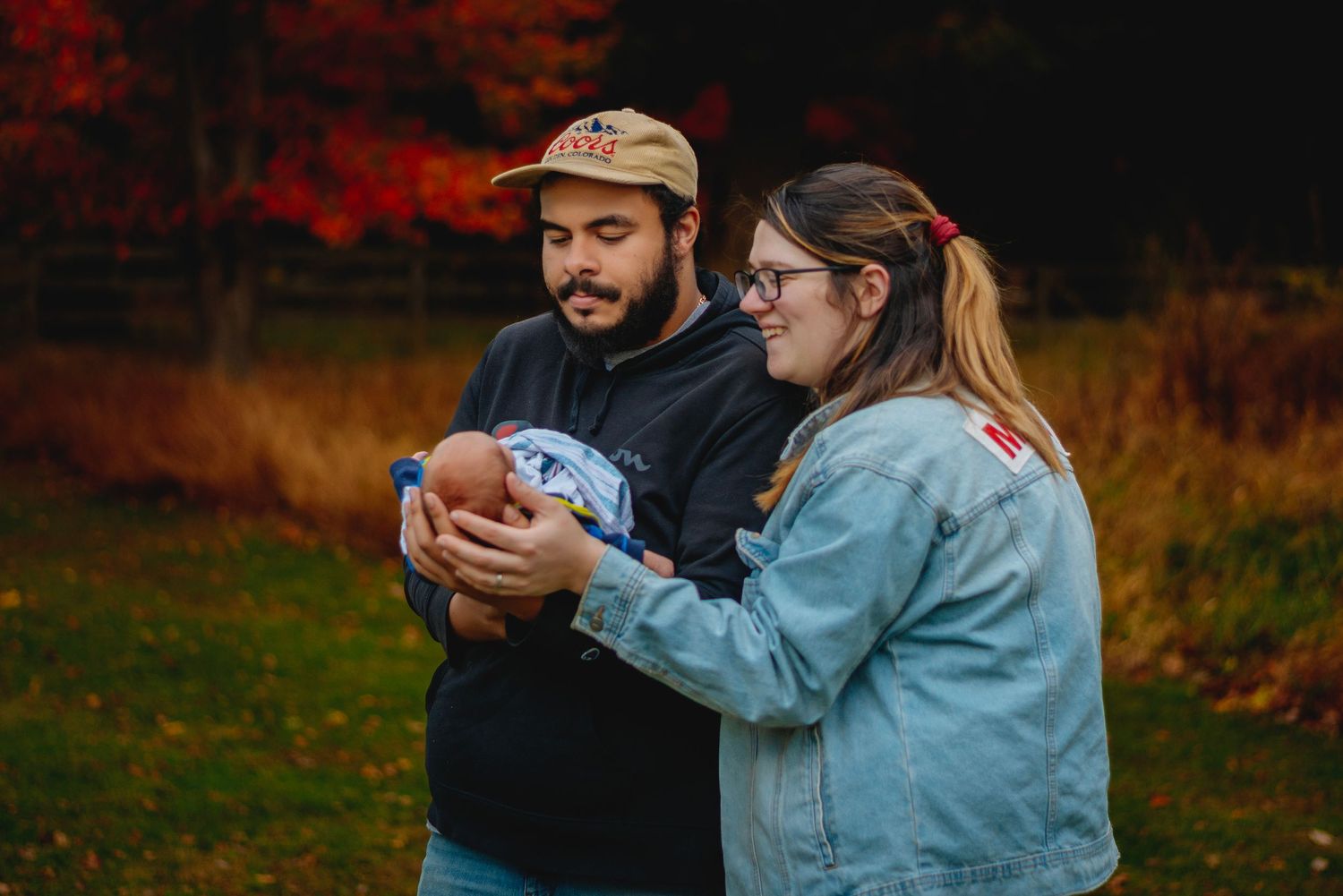 Couple holding newborn baby outdoors in fall with vibrant red foliage in background.
