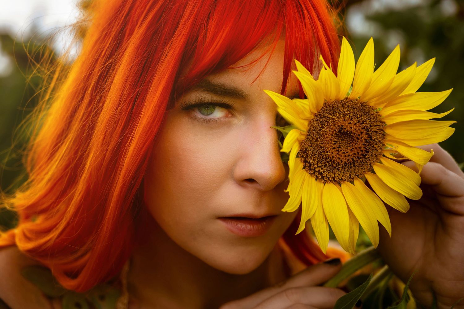A woman with vibrant red hair holds a bright yellow sunflower up to her face outdoors.