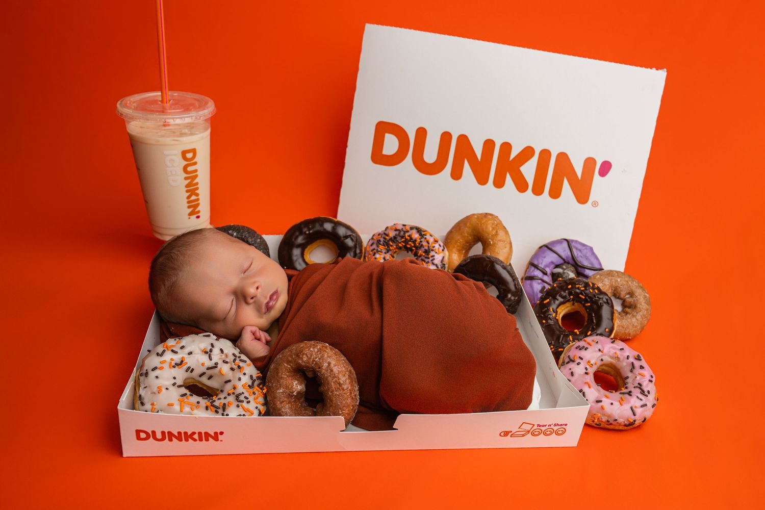 A newborn baby wrapped in brown swaddle sleeps in a Dunkin' donut box surrounded by colorful donuts and iced coffee.