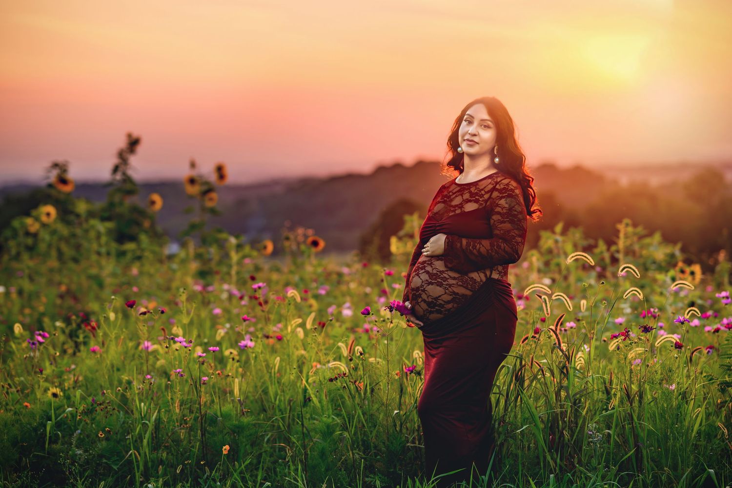 Pregnant woman in red dress standing in wildflower field at sunset.