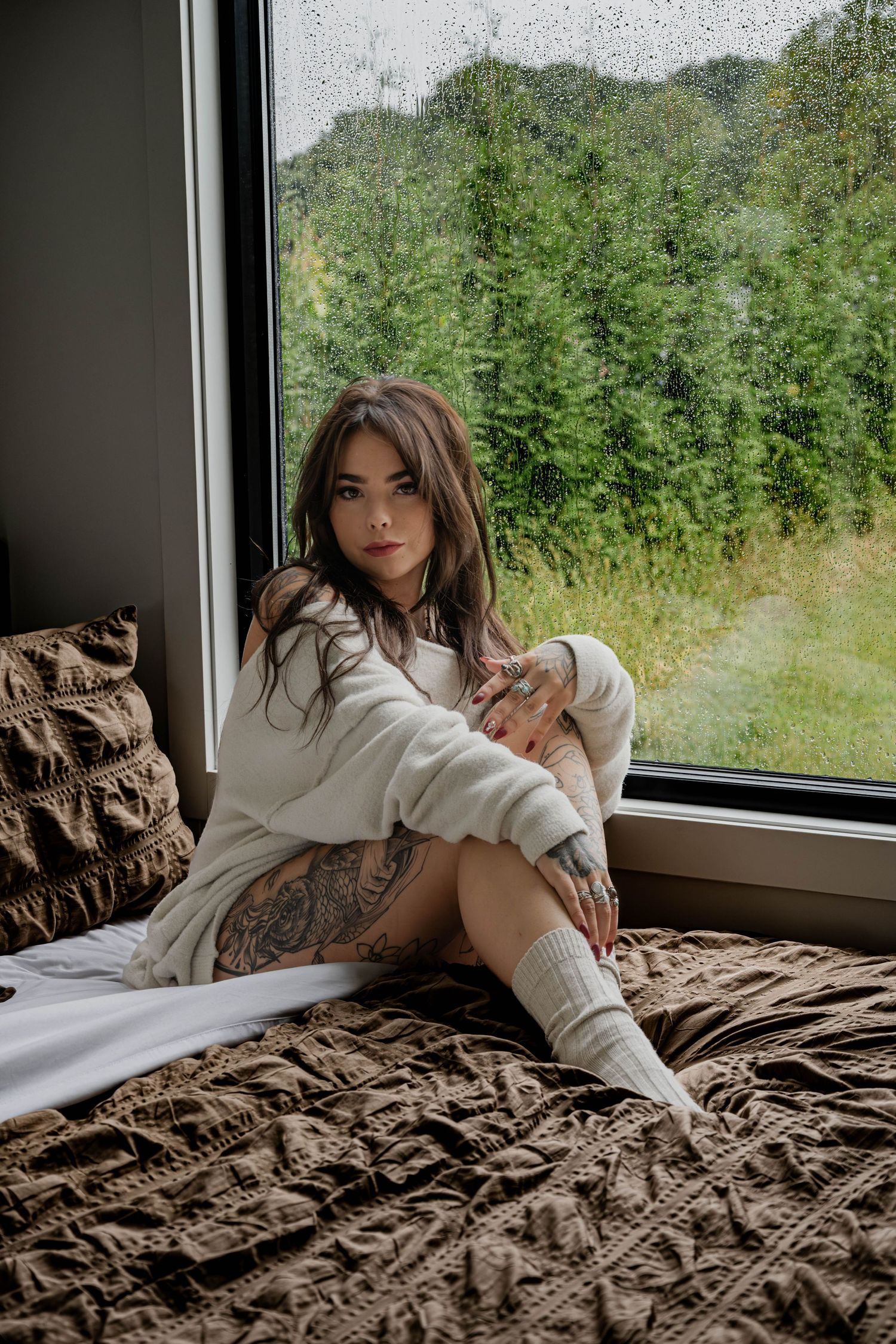 Young tattooed woman in white sweater sitting on bed by rain-covered window with forest view.