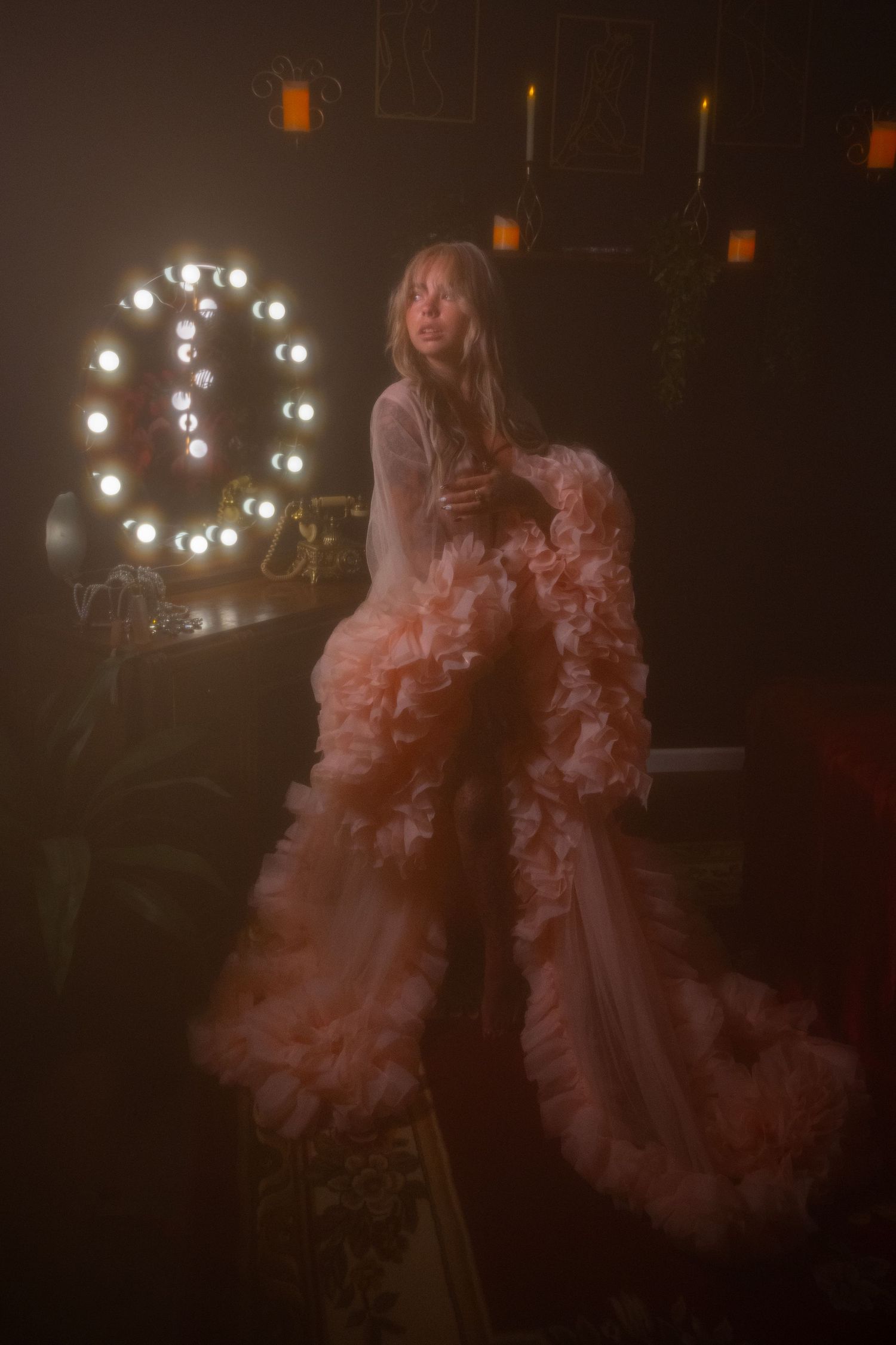 Elegant woman in voluminous peach ruffled gown posing in a dark candlelit room with marquee light.