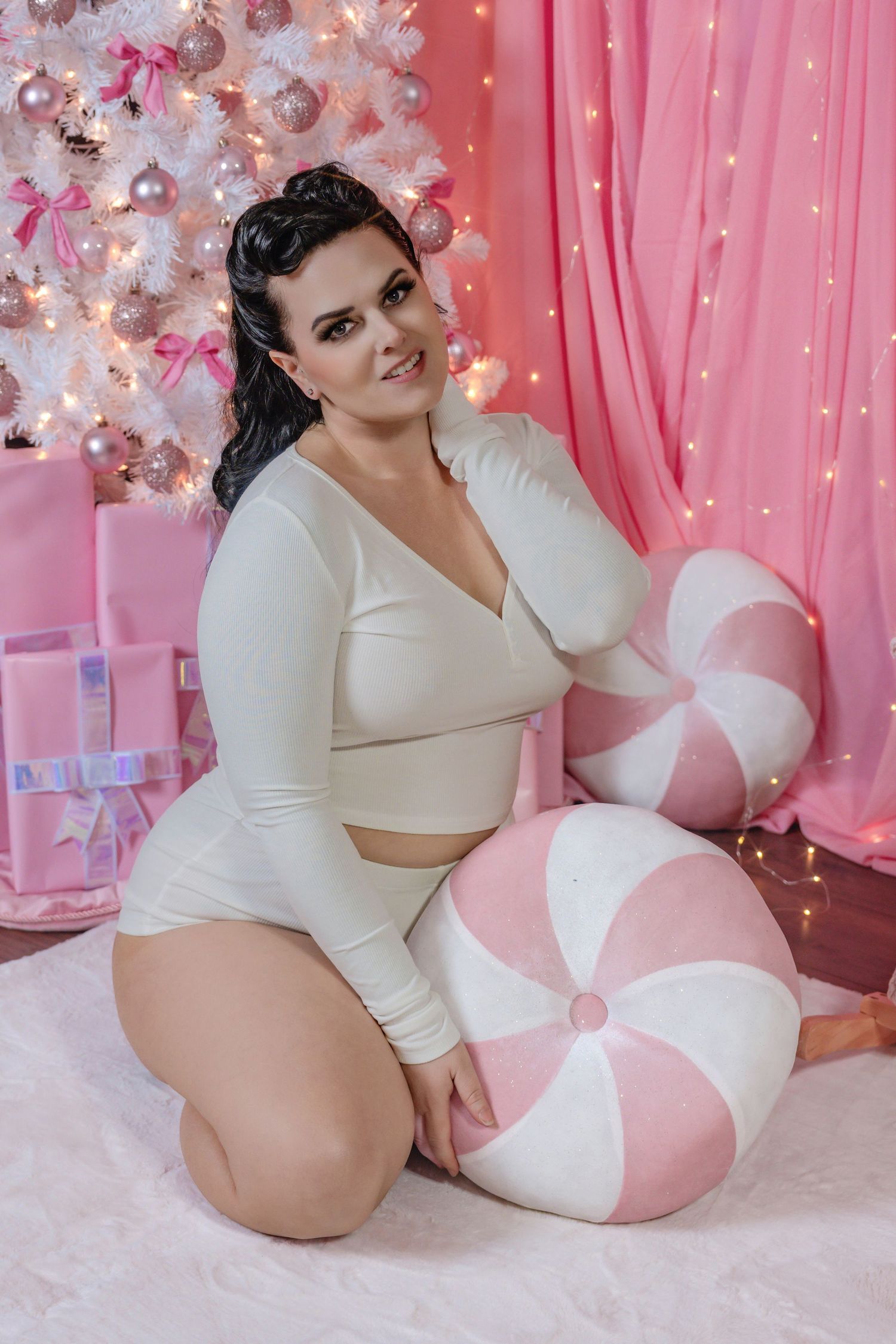 Plus-size brunette woman in cream crop set sitting with large pink candy pillow near Christmas tree.