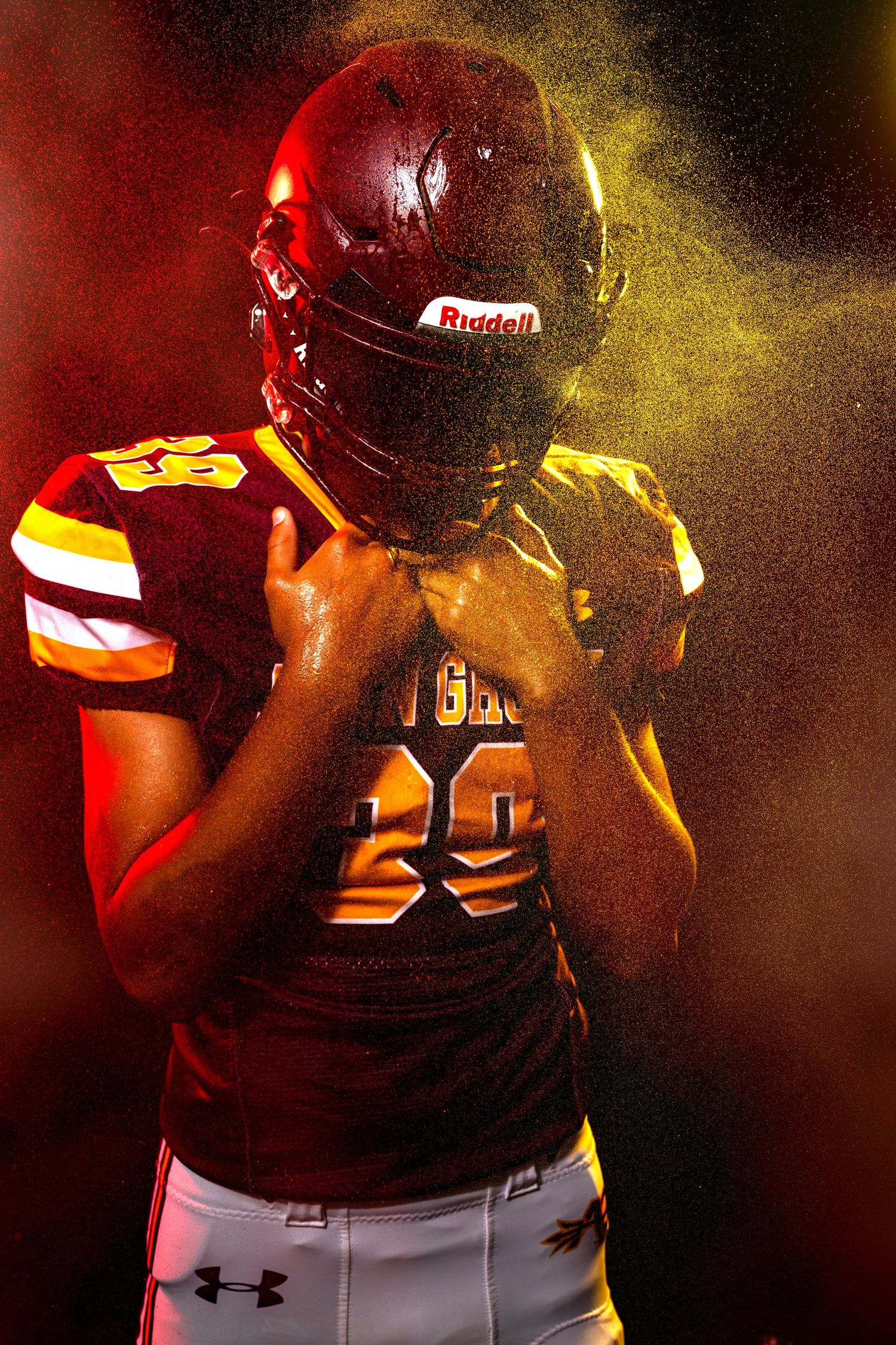 Football player in maroon Riddell helmet and Under Armour uniform posed dramatically with yellow powder explosion.