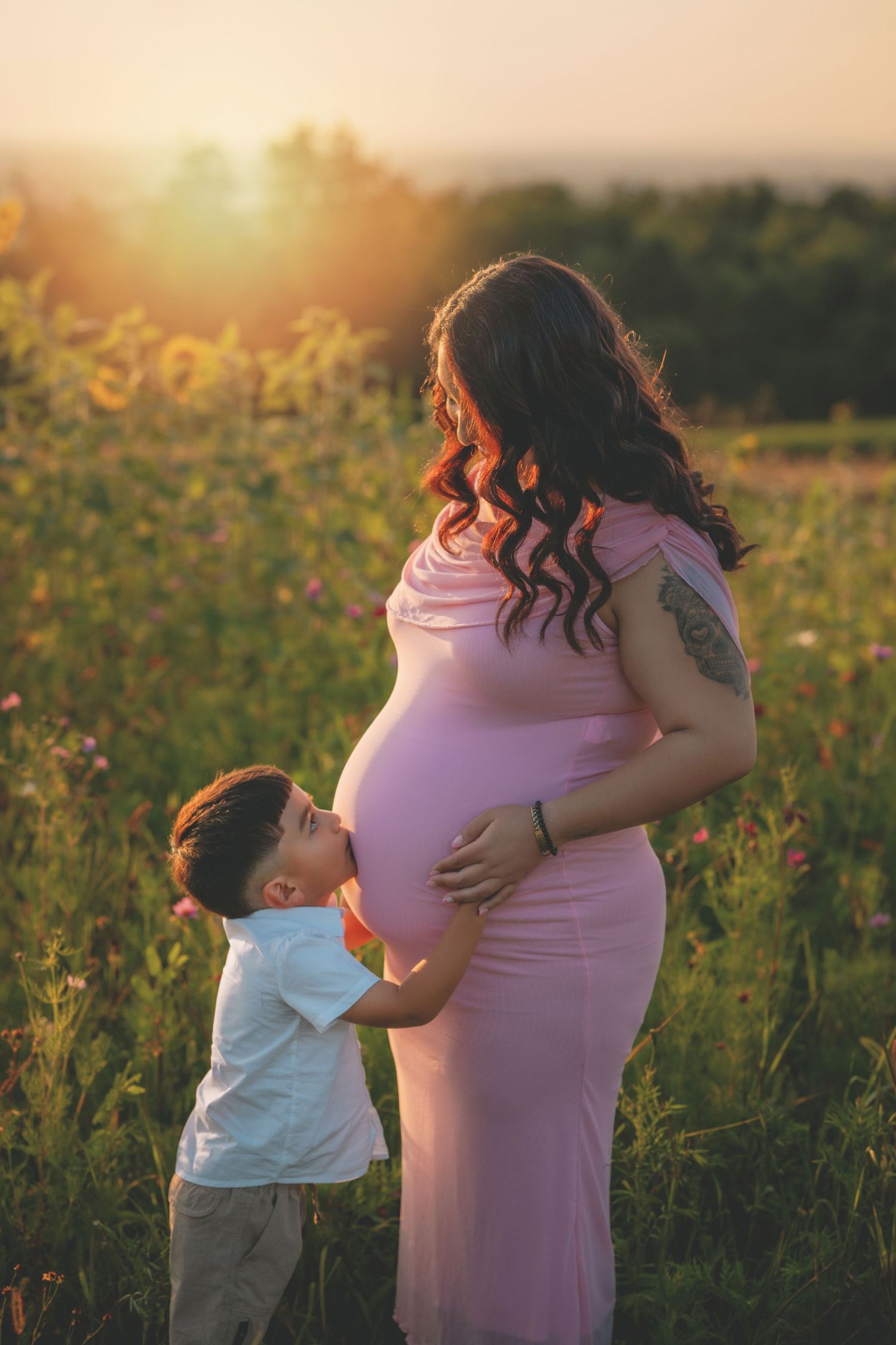 Young boy kissing pregnant mothers belly in a sunlit wildflower field at sunset.