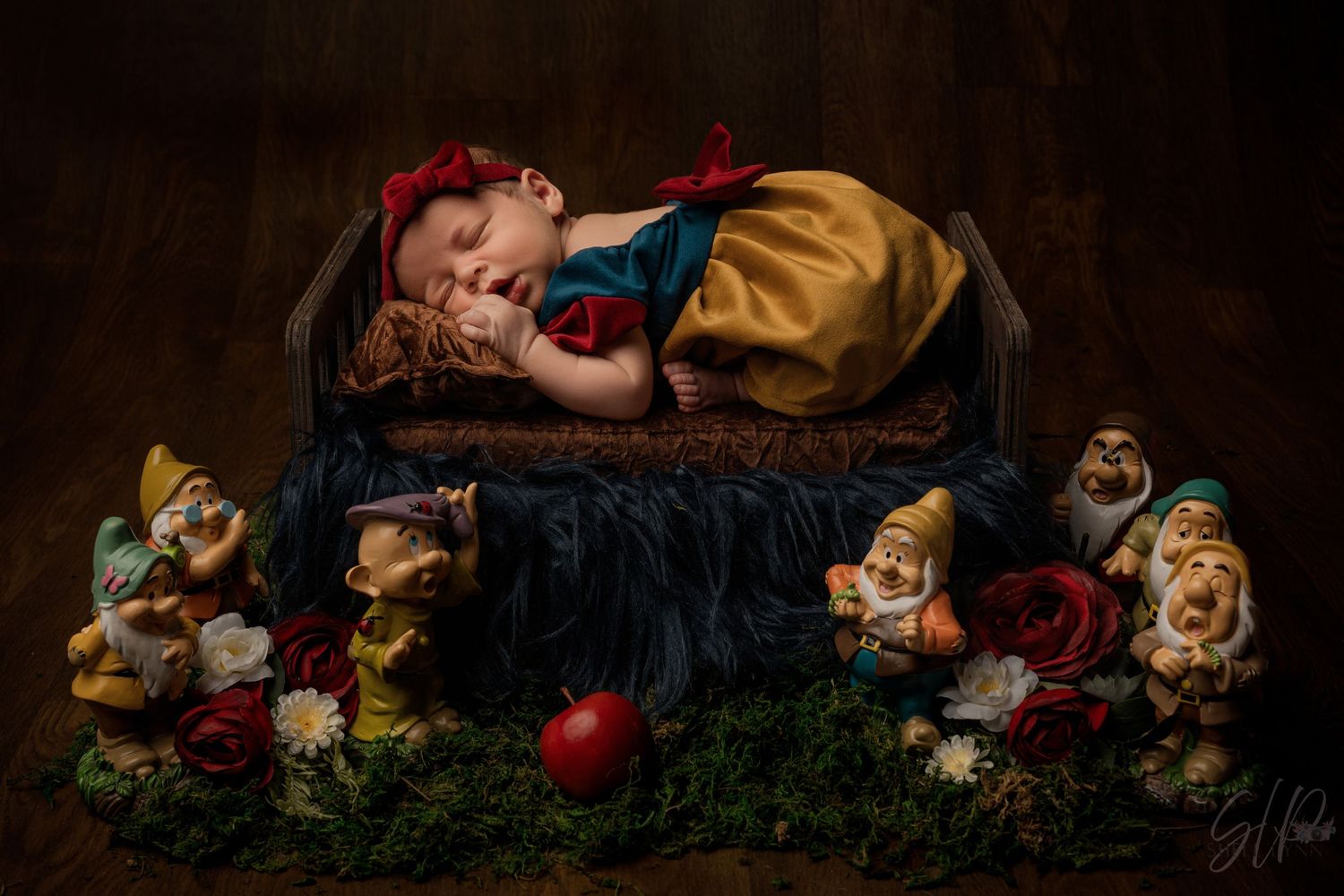 Newborn baby dressed as Snow White sleeping on a tiny bed surrounded by Seven Dwarfs figurines and red roses.