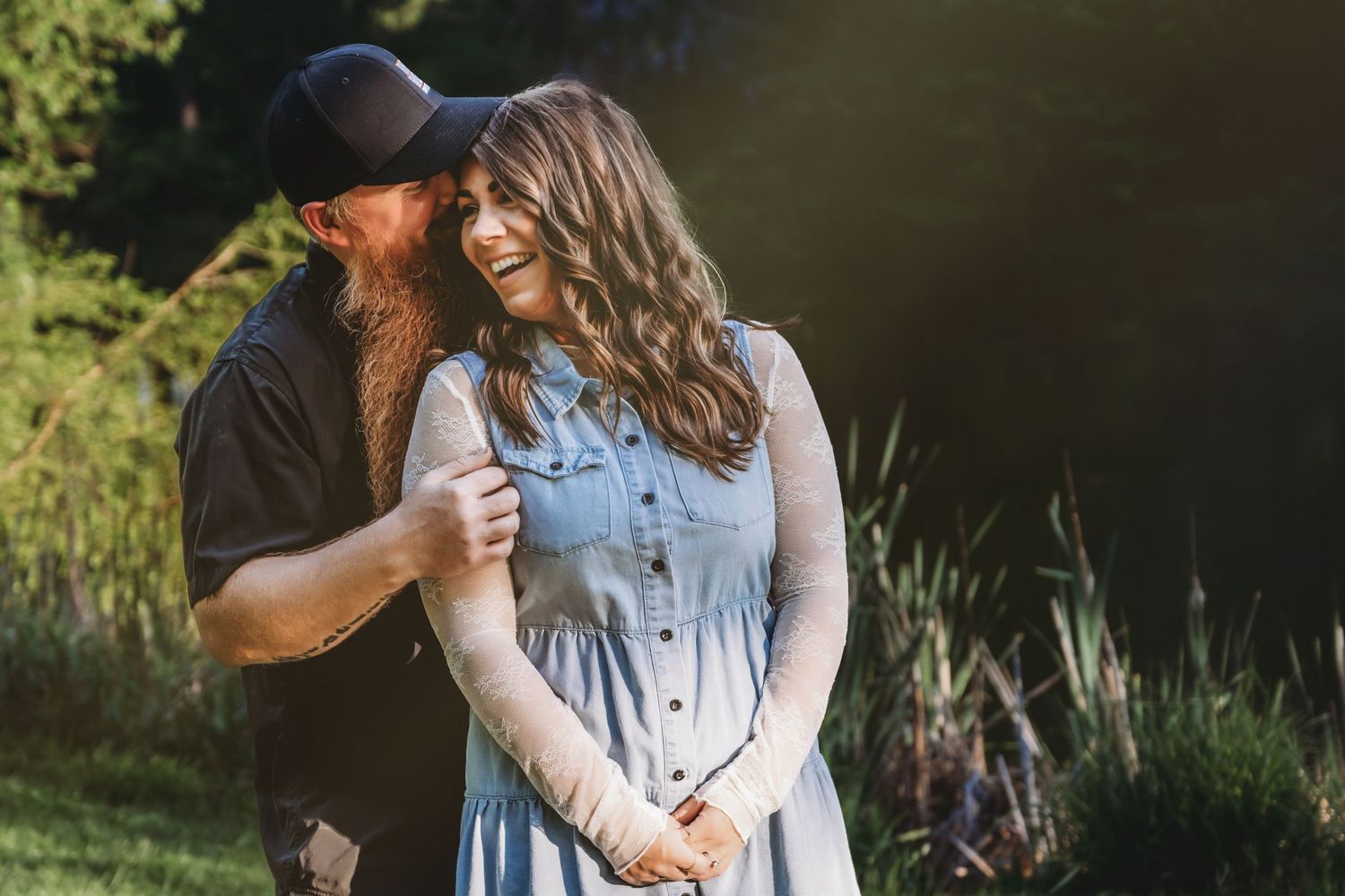 Couple embracing outdoors in golden sunlight, woman in blue dress and man in black shirt and cap, smiling together.