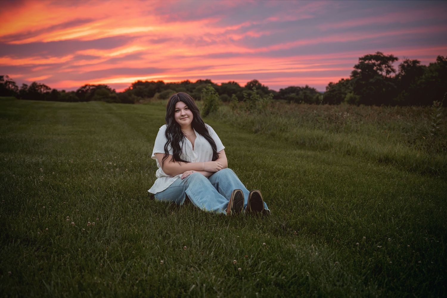 Young woman sitting in green grass field during vibrant pink and orange sunset.