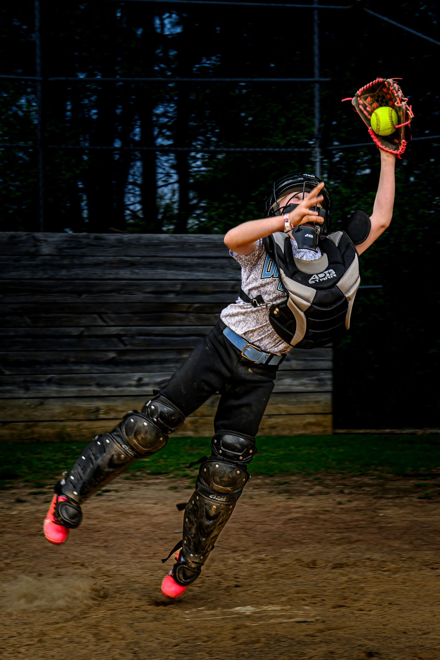 Young softball catcher in full gear leaping to catch a yellow ball during a game on a dirt field.