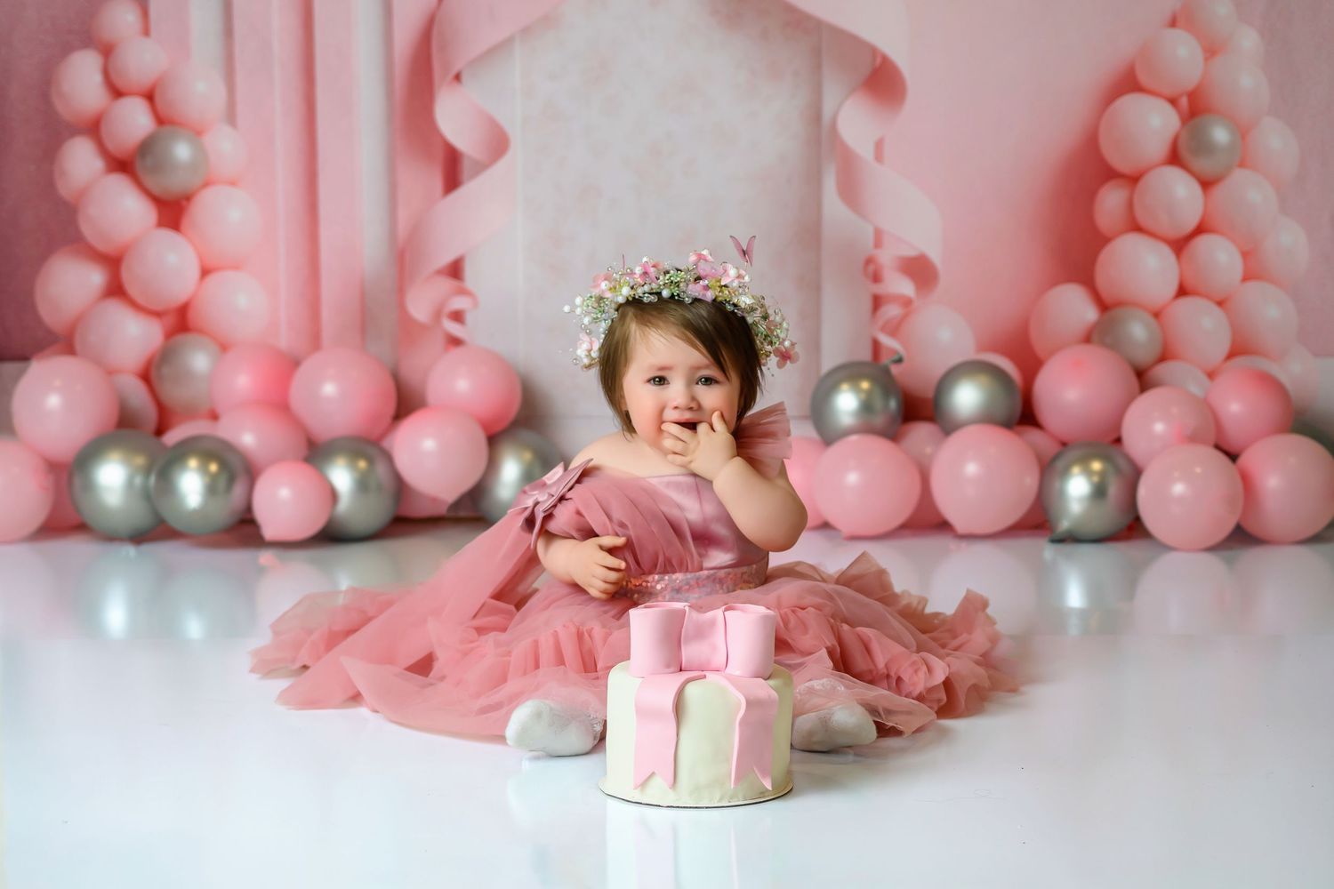 Sweet baby girl in pink dress with floral crown sitting beside pink birthday smash cake.