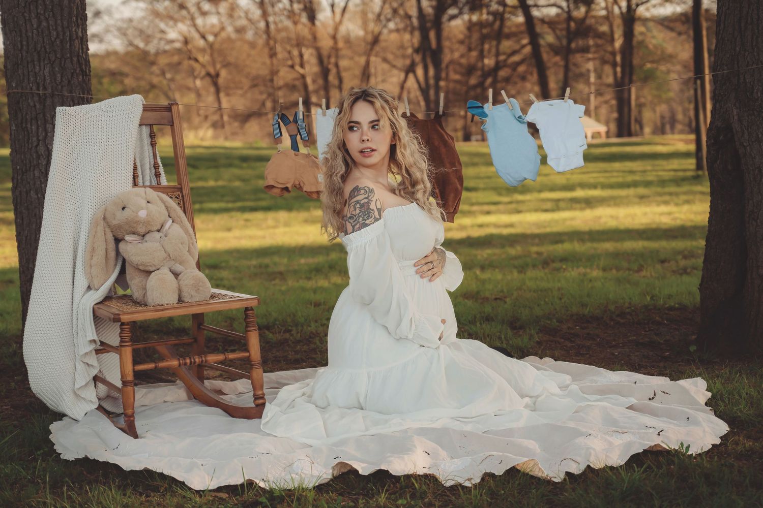 Pregnant woman in white dress sits outdoors near stuffed bunny and blue baby clothes hanging on line.