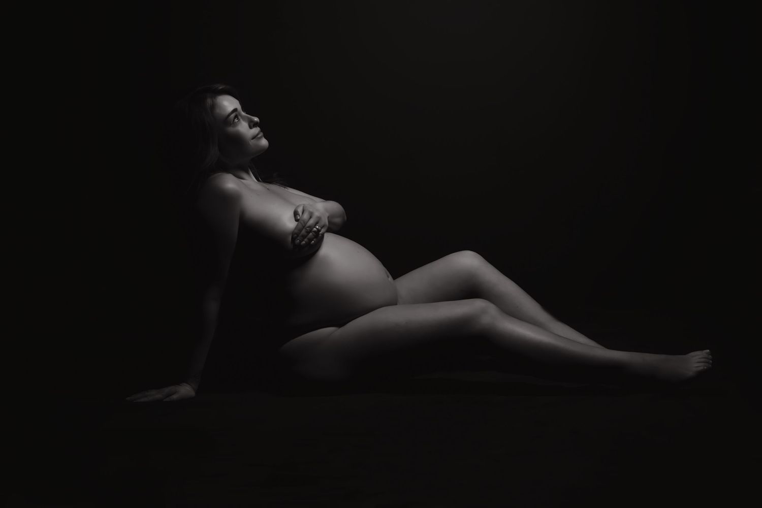 Black and white maternity photo of pregnant woman reclining gracefully against dark background.