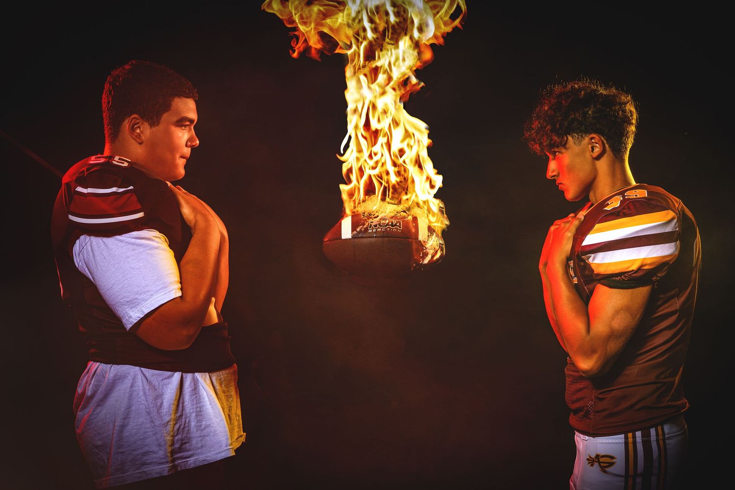 Two football players in maroon uniforms stand facing each other with a football engulfed in flames.