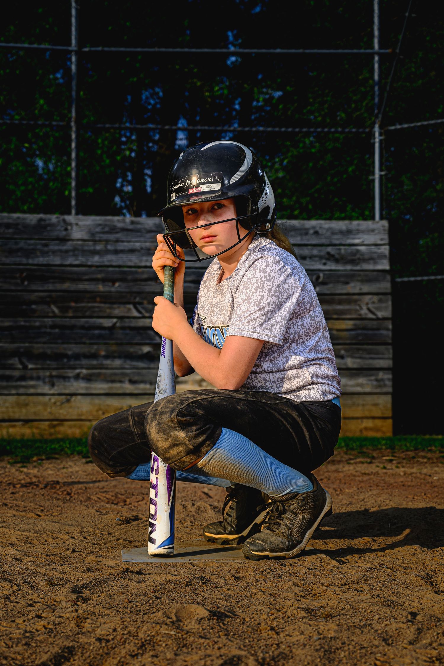 Young softball player kneels with bat on dirt field wearing black helmet at dusk.