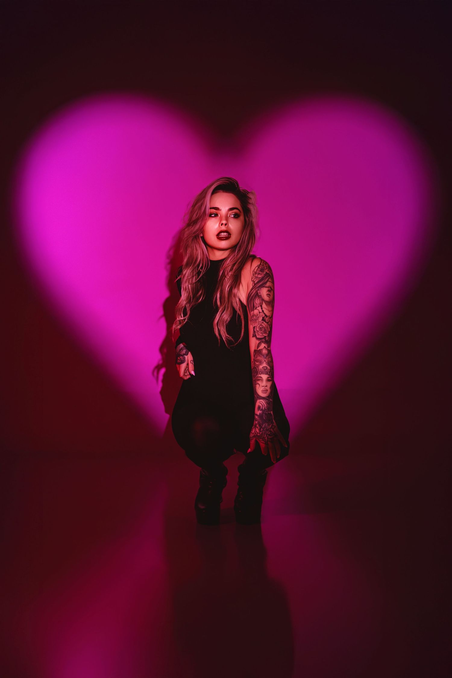 Tattooed woman in black outfit poses before glowing pink heart-shaped light on dark background.