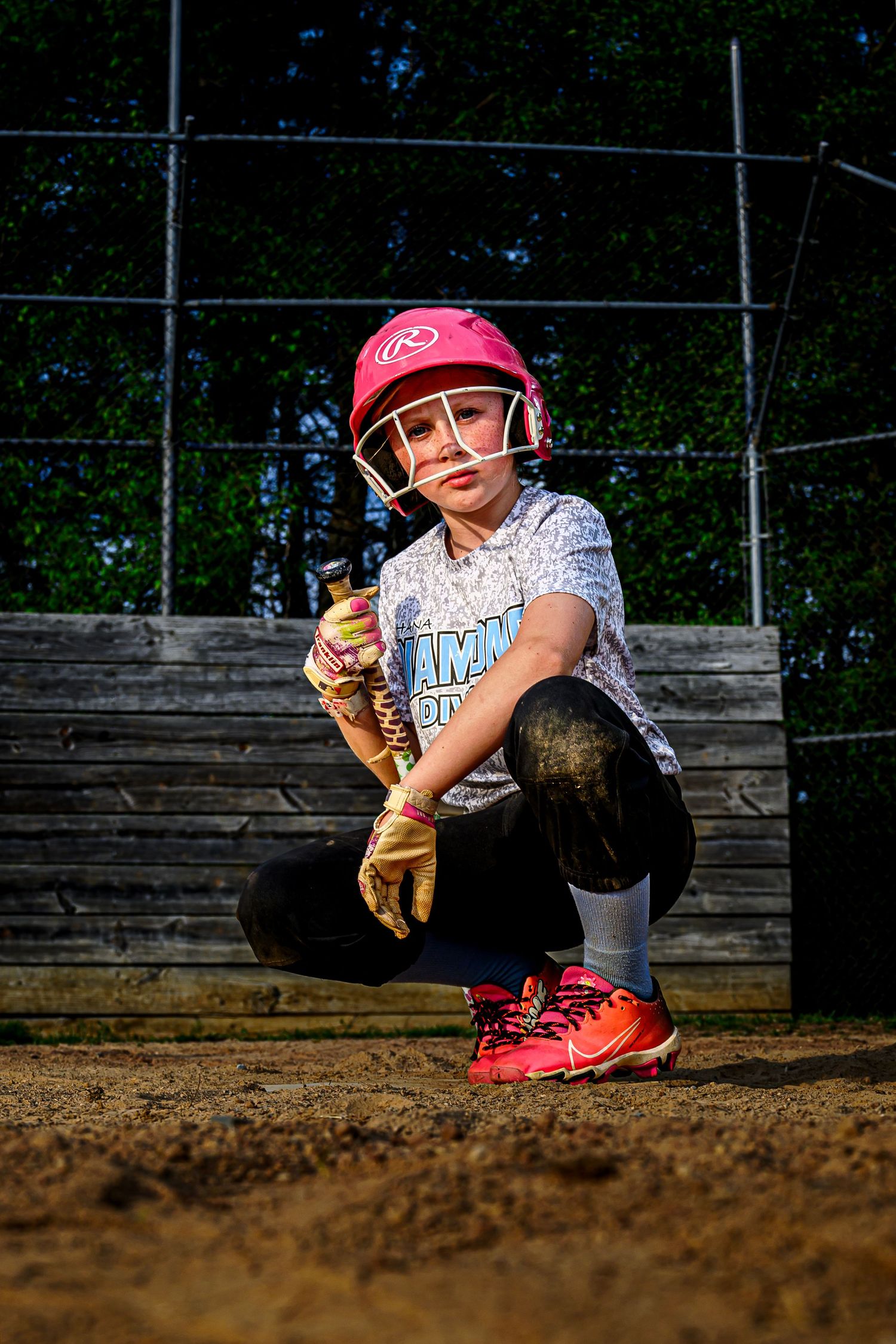Young girl softball player crouches in pink helmet and batting gloves on dirt field.