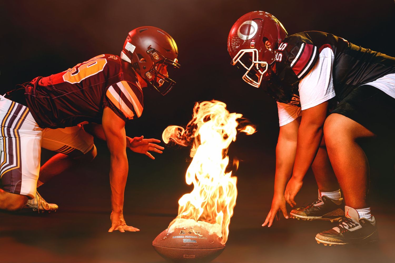 Two football players in maroon uniforms face off over a flaming football on the ground.