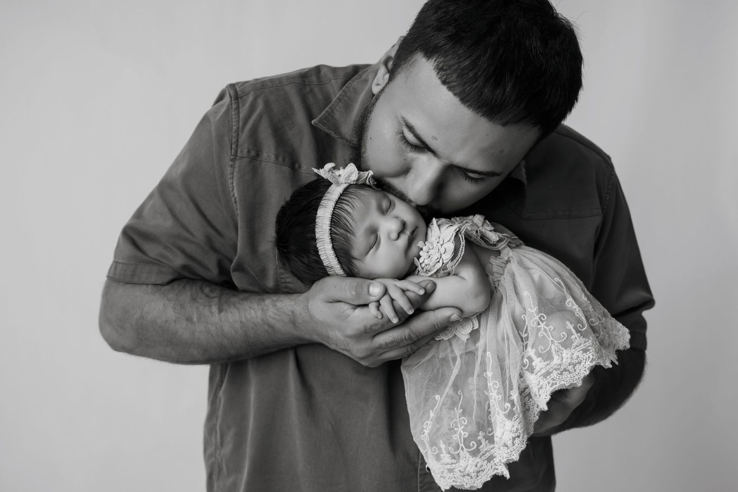 Black and white photo of a father tenderly kissing his newborn baby girl in a lace outfit.