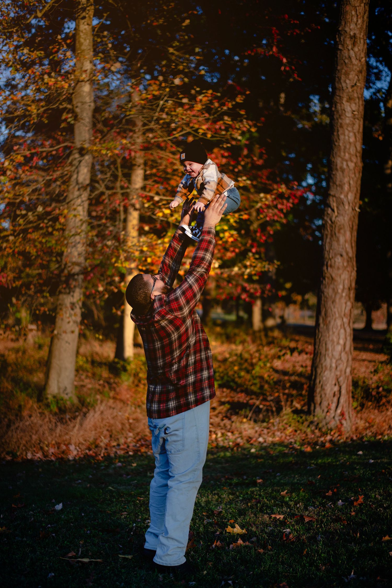 Father lifting baby overhead in autumn park, wearing plaid shirt and jeans among colorful fall foliage.