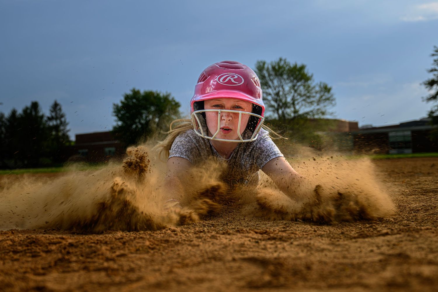 Female softball player in pink Rawlings helmet dives headfirst into base in cloud of dust.