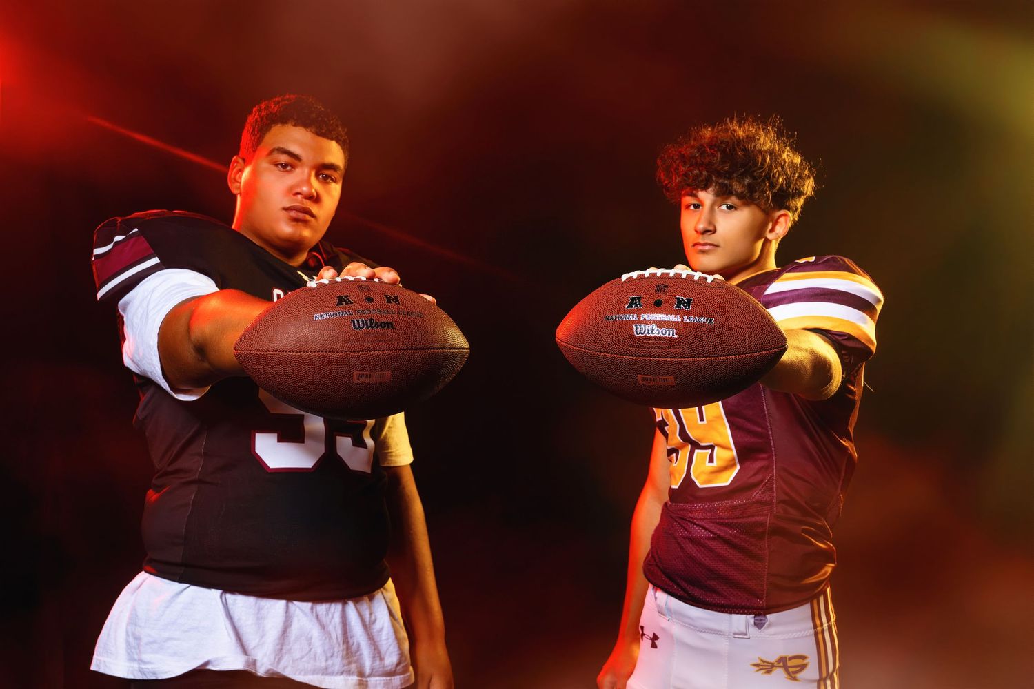 Two Avon Grove football players in maroon uniforms extend footballs toward camera in dramatic pose.