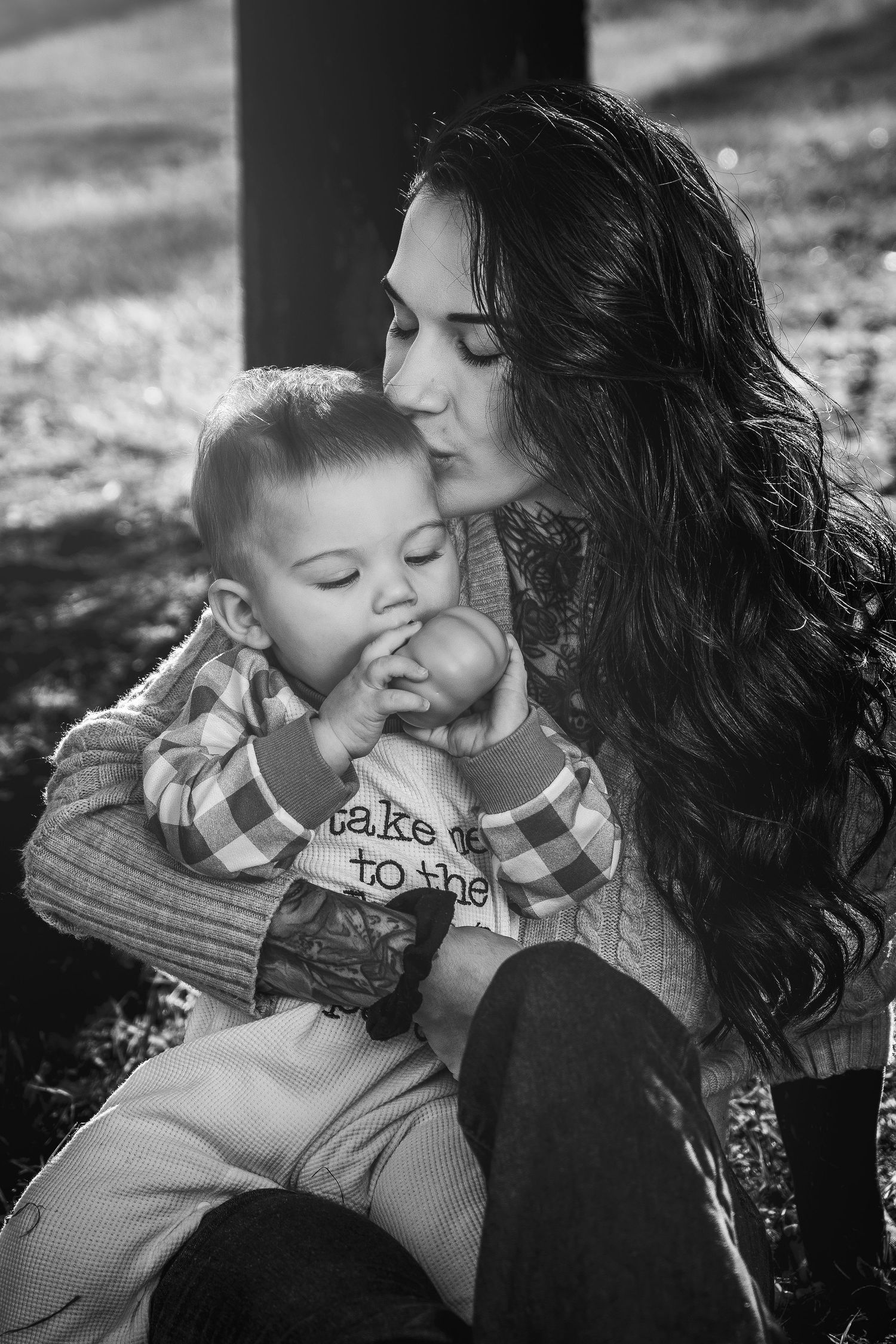 Black and white photo of a loving mother kissing her baby boy on the cheek while he sucks his thumb outdoors.