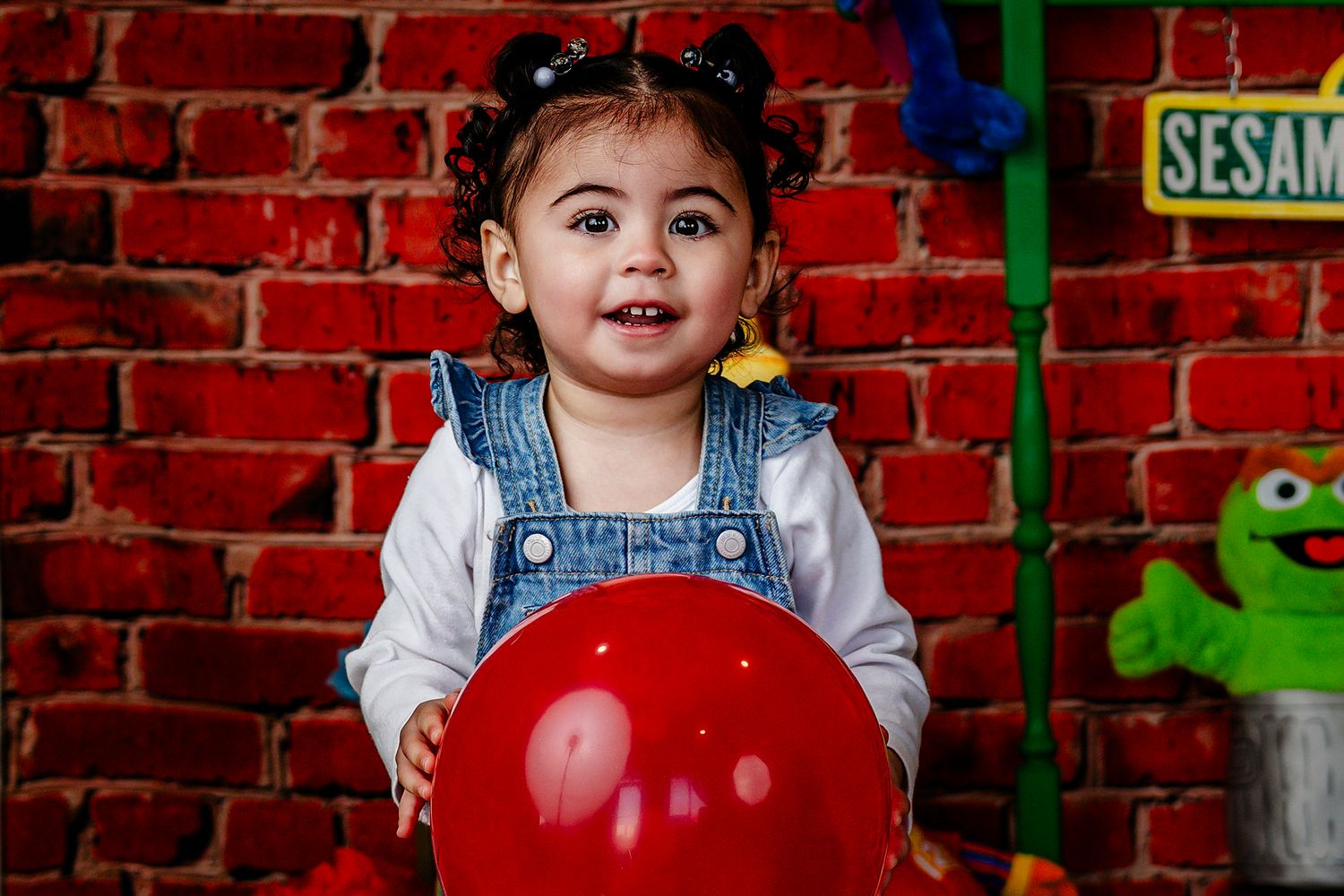 Smiling toddler girl in denim overalls holds red balloon before brick wall backdrop.