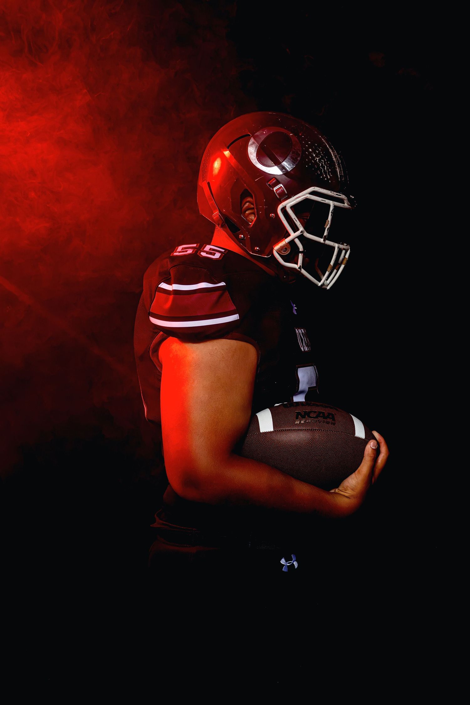 High school football player in red helmet holds ball in dramatic dark smoky side profile.