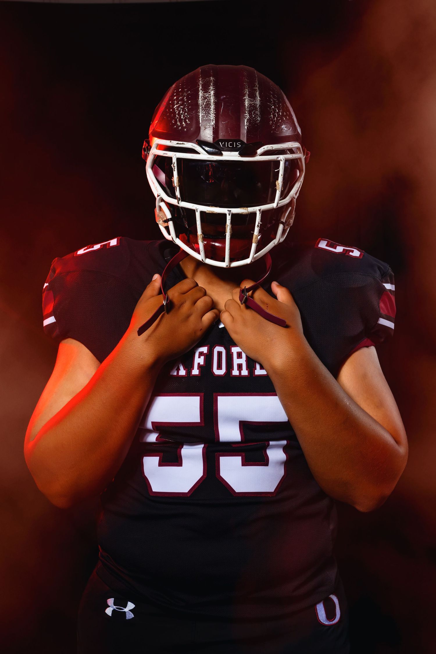 Oxford #55 football player adjusts helmet chin strap in dramatic red foggy studio shot.