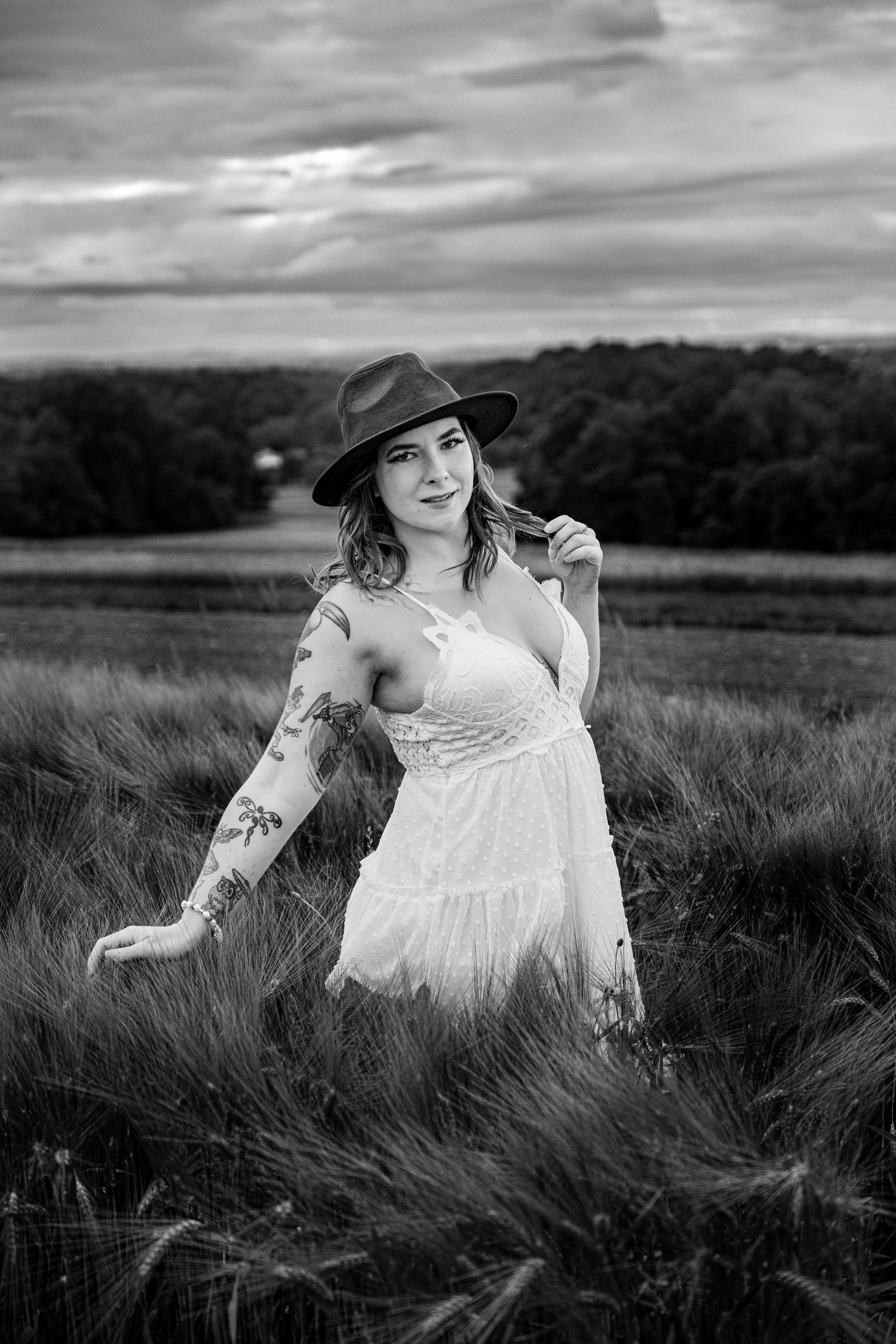 Black and white portrait of tattooed woman in white dress and hat standing in a grain field.