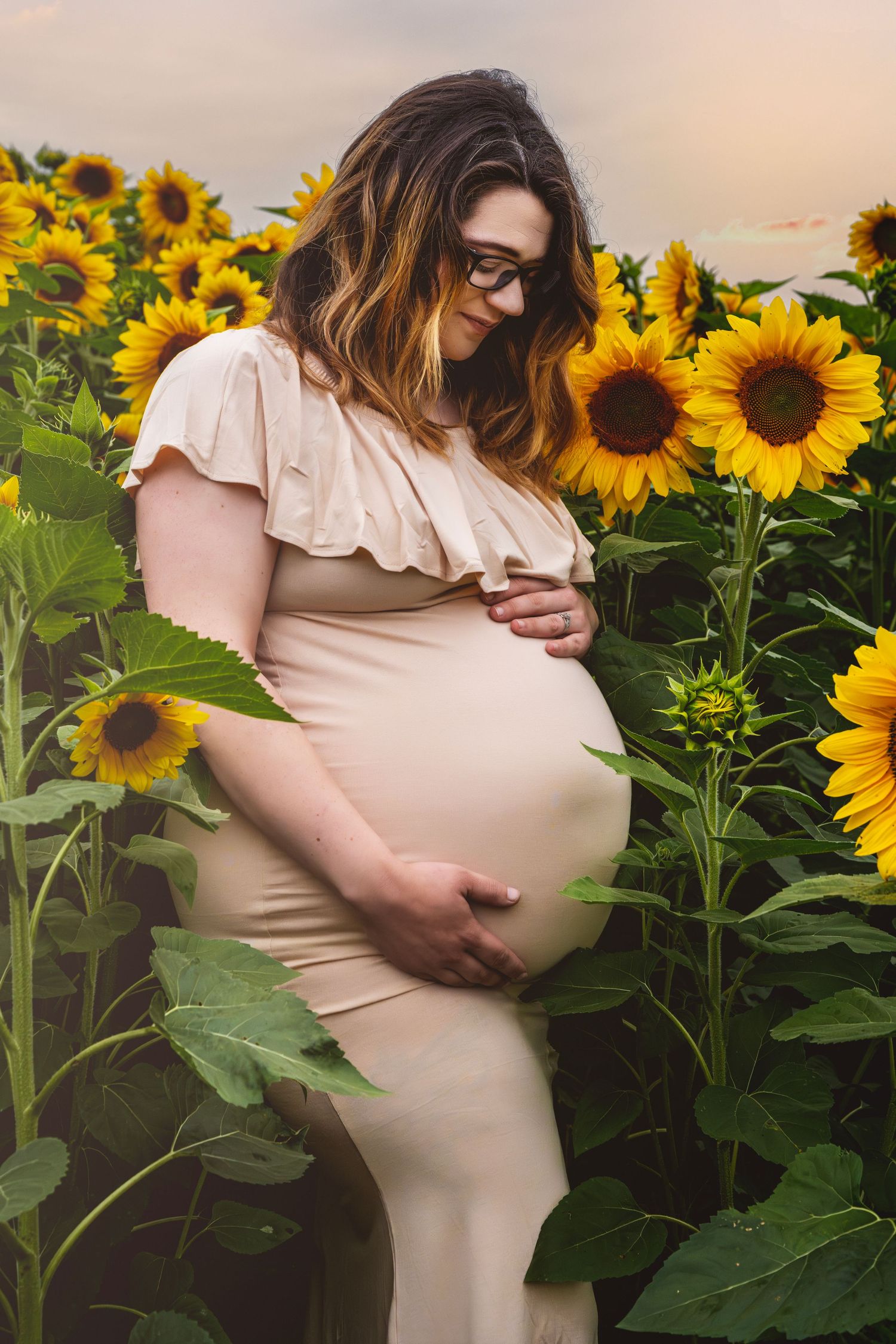Pregnant woman in cream dress cradling her baby bump in a vibrant sunflower field at sunset.