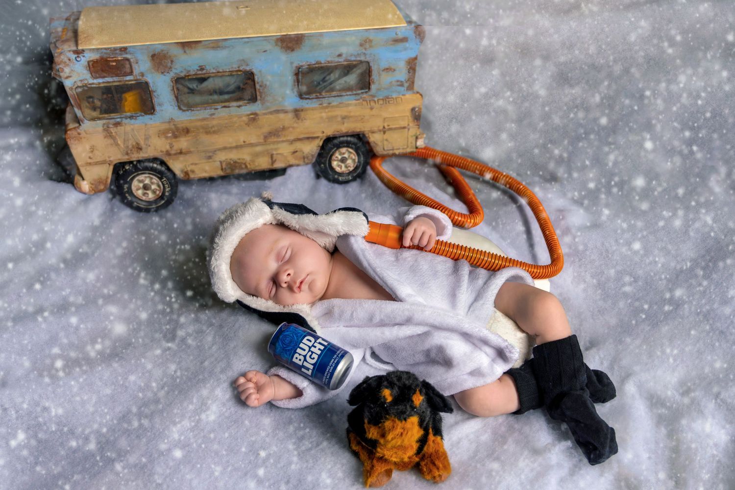 Newborn baby sleeping in white robe with hat beside toy bus and stuffed dog on snowy background. Cousin Eddie newborn photo