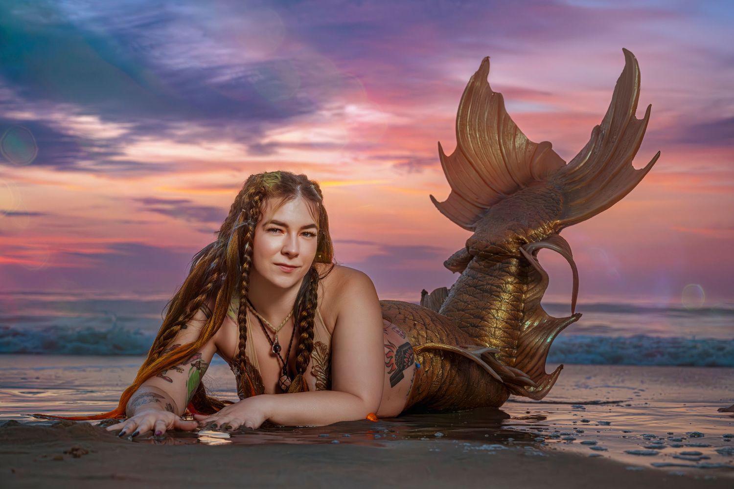 Woman dressed as a mermaid with golden tail poses on sandy beach at colorful sunset.