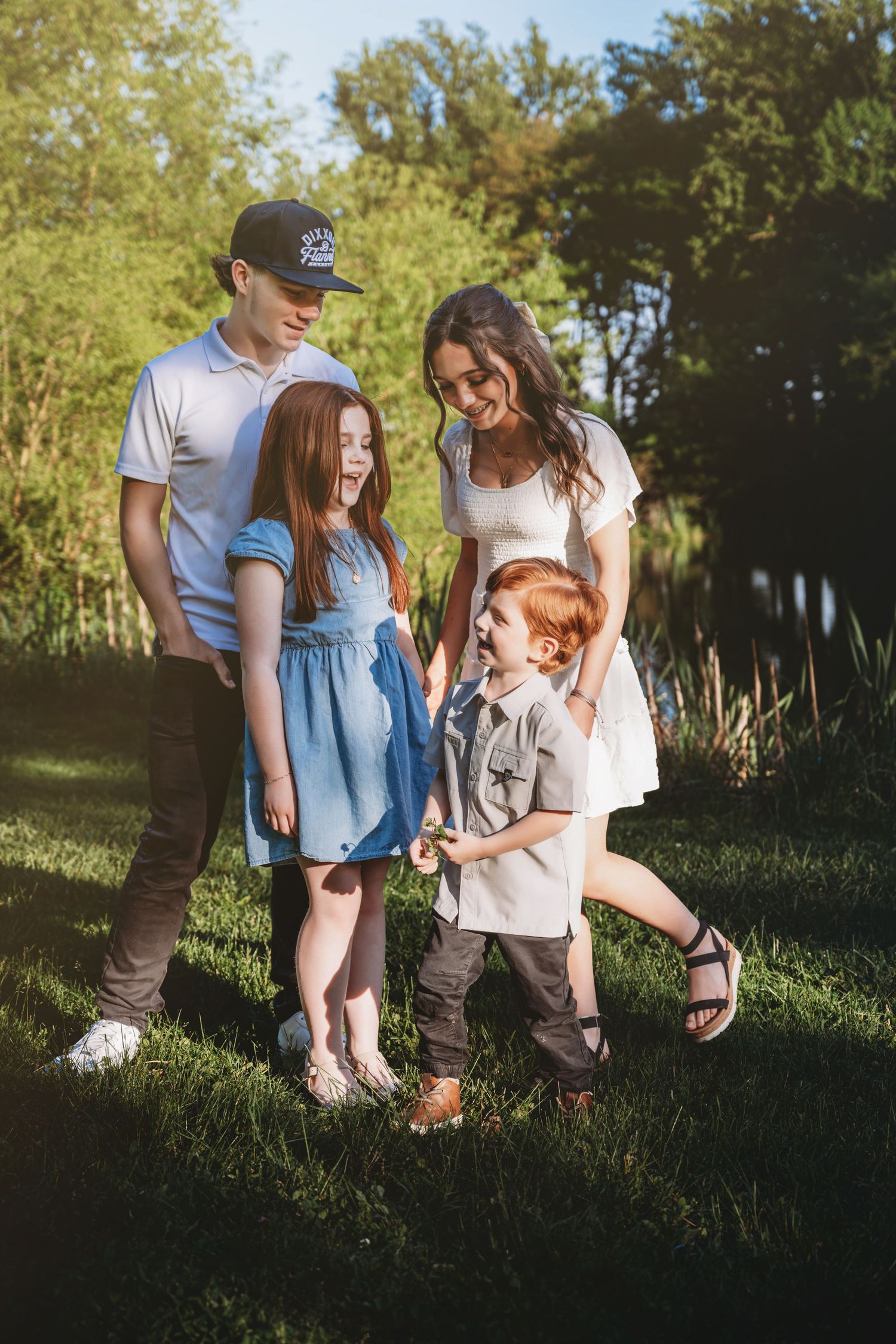 A happy family of four enjoys a sunny outdoor moment on green grass, with two redhead children laughing together.