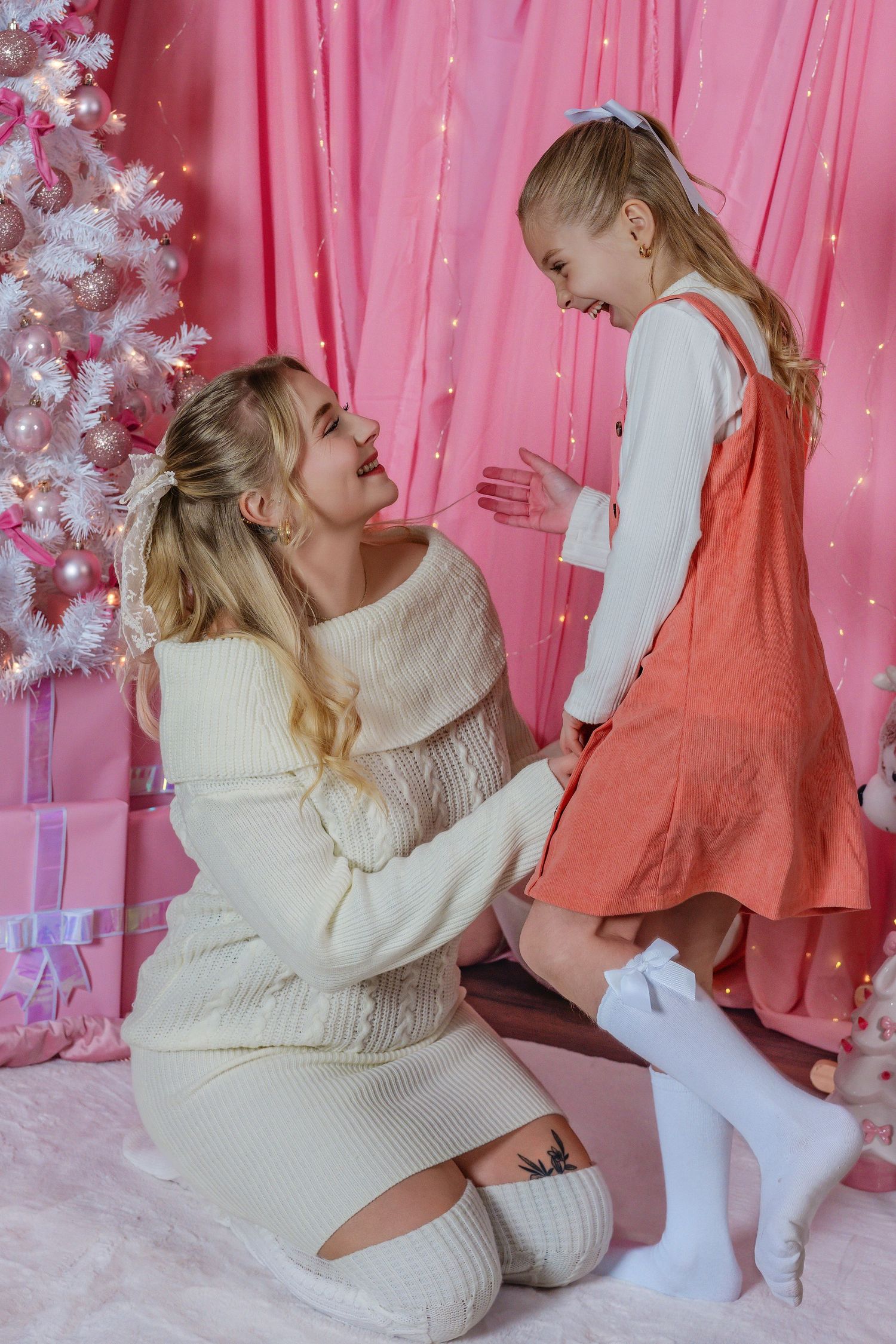 A mother kneels beside her smiling daughter near a white Christmas tree in a pink holiday decorated room.