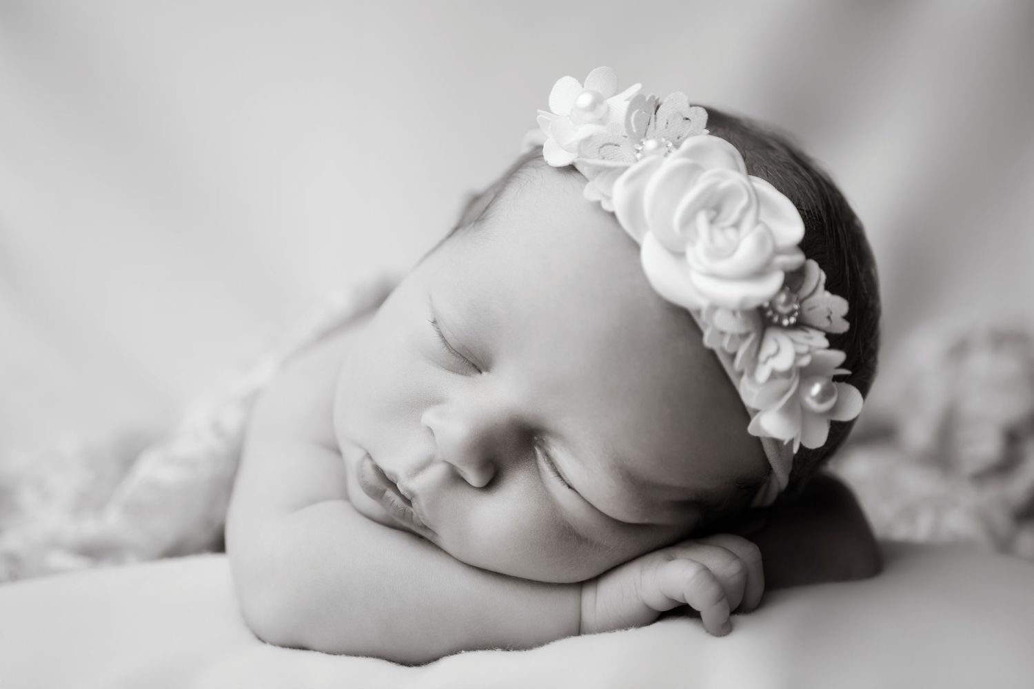 Black and white newborn photo of sleeping baby girl wearing white floral headband, peacefully resting on hands.